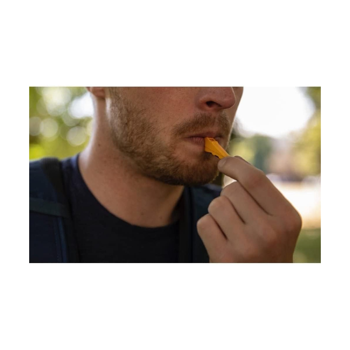 Ein Mann mit Bart bläst in eine gelbe Trillerpfeife. Bäume im Hintergrund.