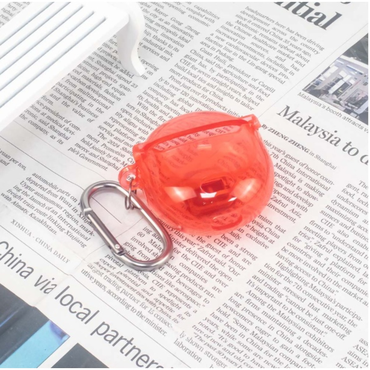 Ein roter, durchsichtiger Ohrhörerbeutel mit Karabiner liegt auf einer Zeitung.