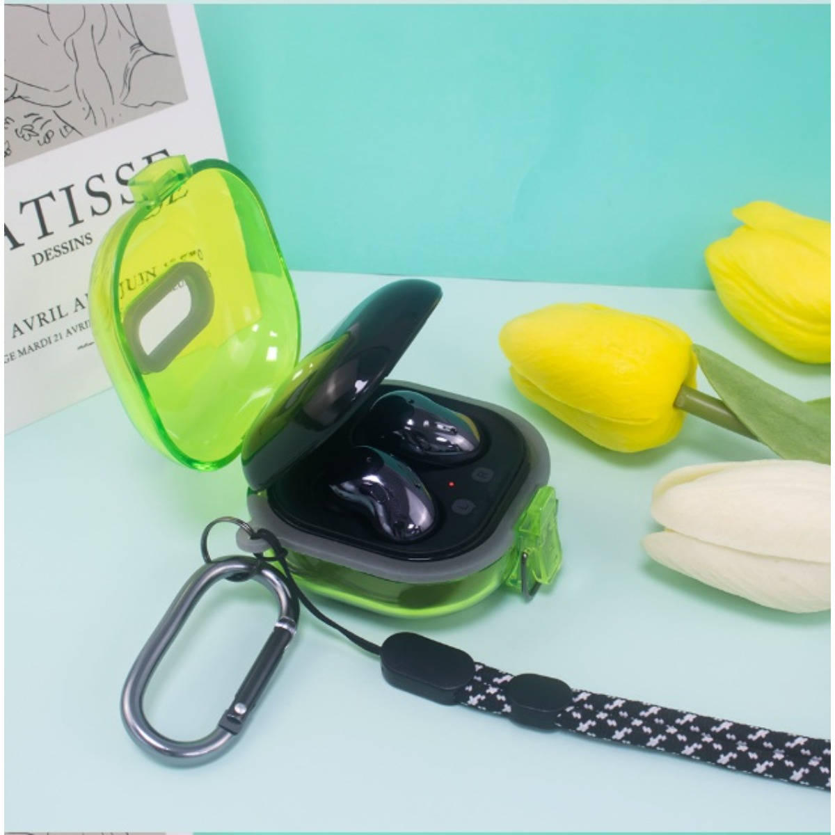 Schwarze kabellose Ohrhörer in einem durchsichtigen grünen Etui mit Schlaufe neben gelben Tulpen.