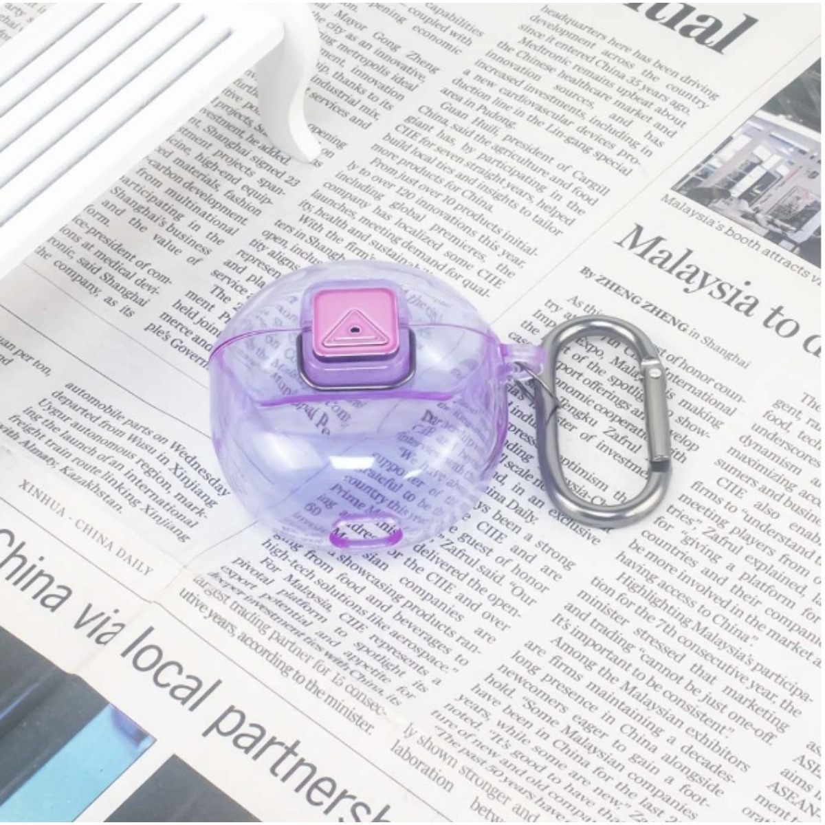 Una custodia viola traslucida per auricolari con chiusura rosa appoggiata su pagine di giornale.