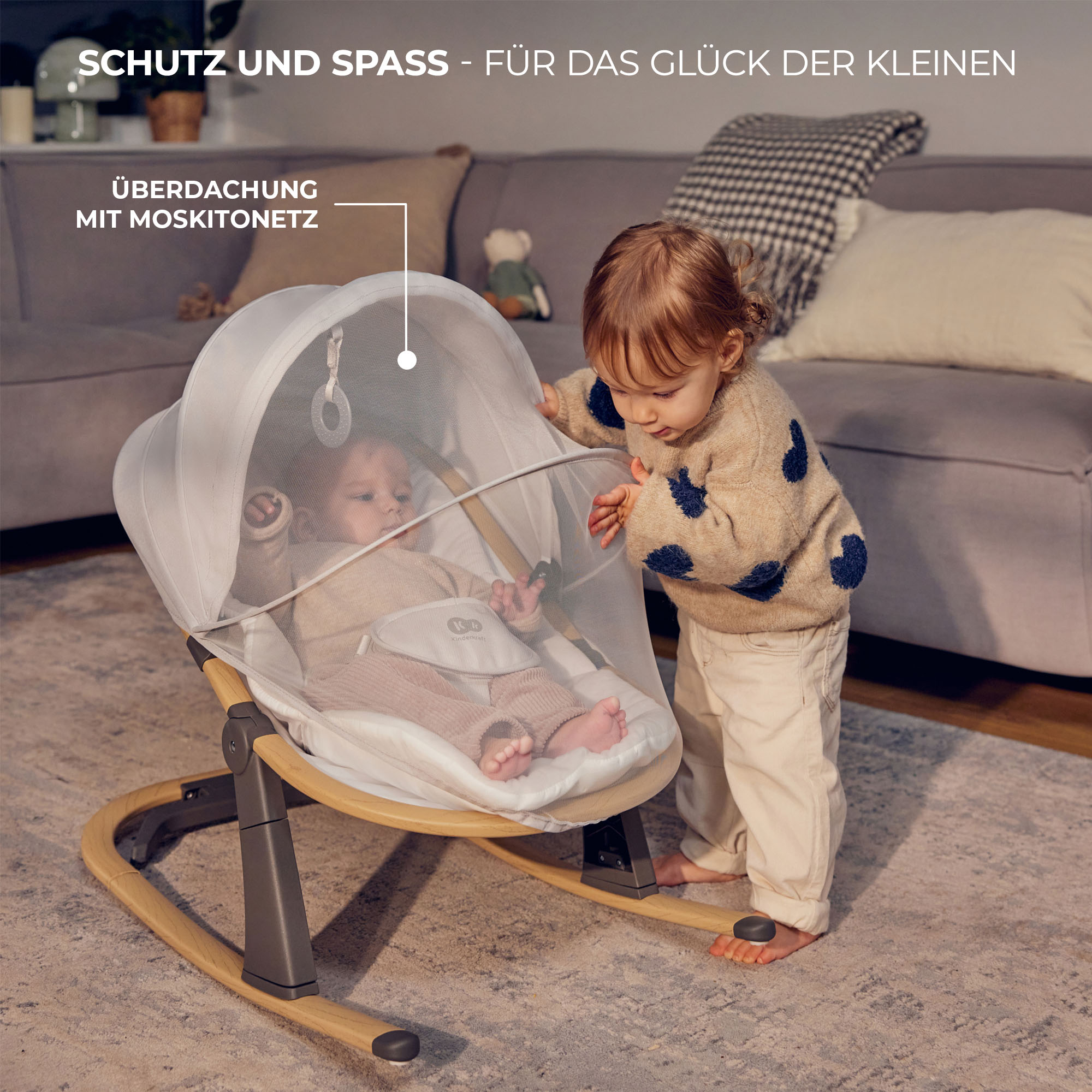 Zwei Kinder in einem Wohnzimmer, eines in einer Babywippe, das andere steht daneben.