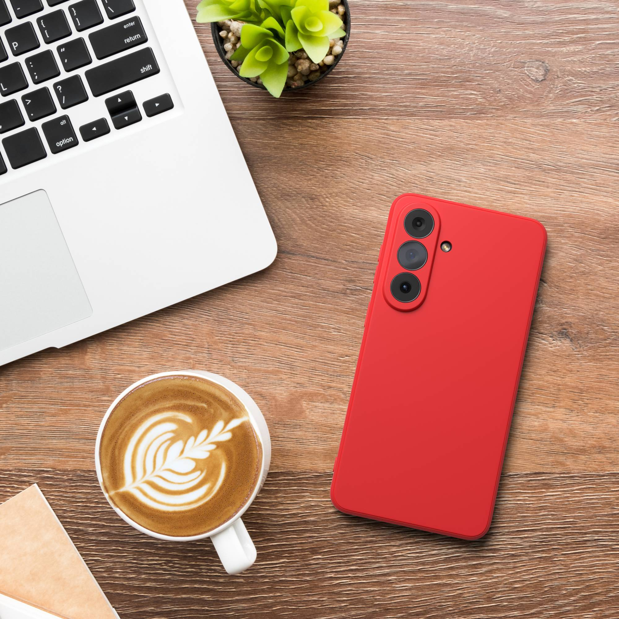 Ein Laptop, eine rote Handyhülle, eine Kaffeetasse und eine Pflanze auf einem Holztisch.