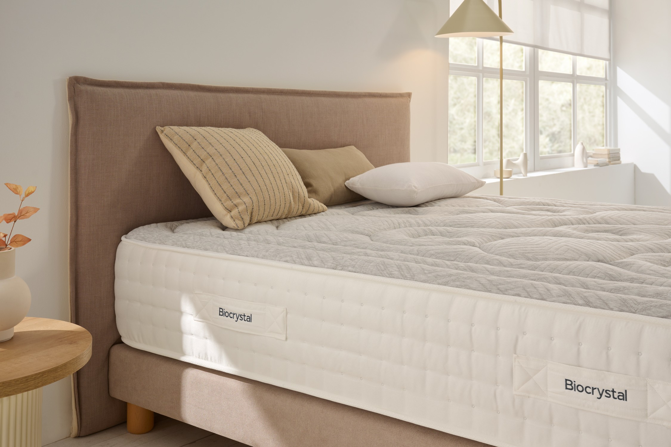 Una cama con cabecero marrón, cojines blancos y colchón Biocrystal.