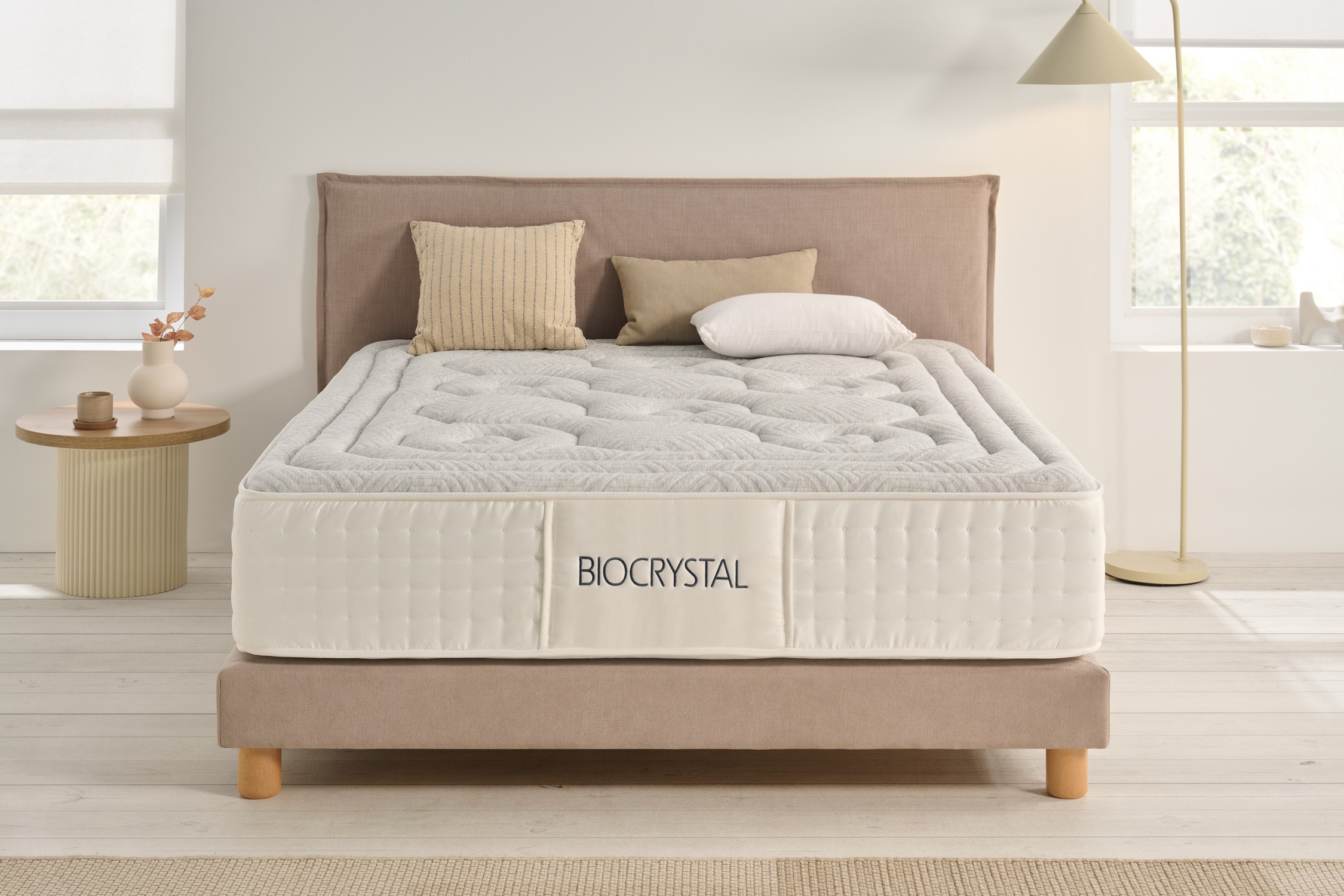 Una cama con colchón blanco y logo Biocrystal, cabecero beige, almohadas.
