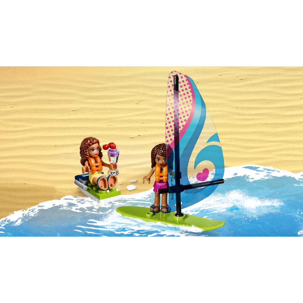 Due figure Lego Friends su una spiaggia sabbiosa con onde blu. Una fa windsurf, l'altra è seduta su una barca con un gelato.