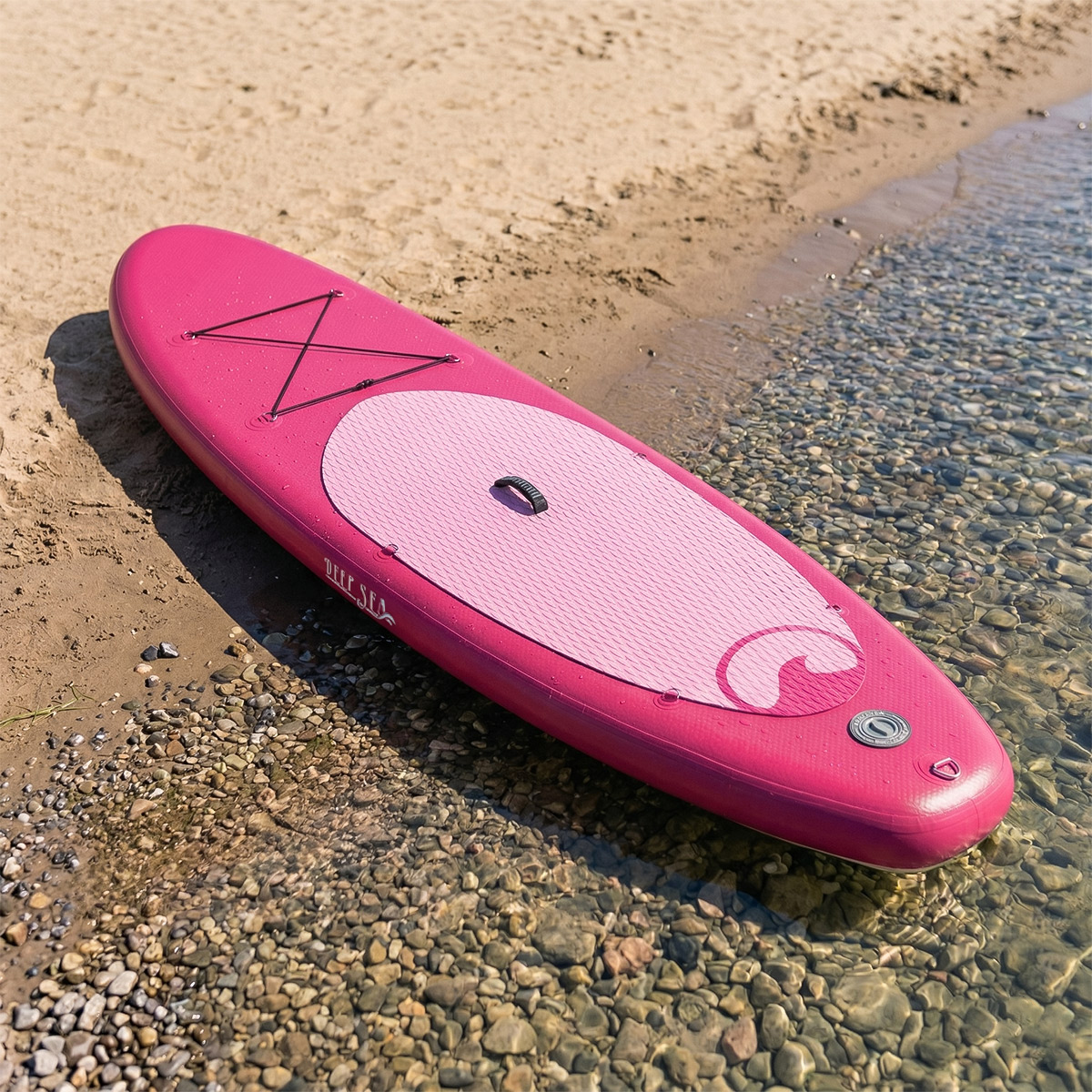 Ein pinkes Paddleboard liegt an einem Kiesstrand mit sanften Wellen und Sand.