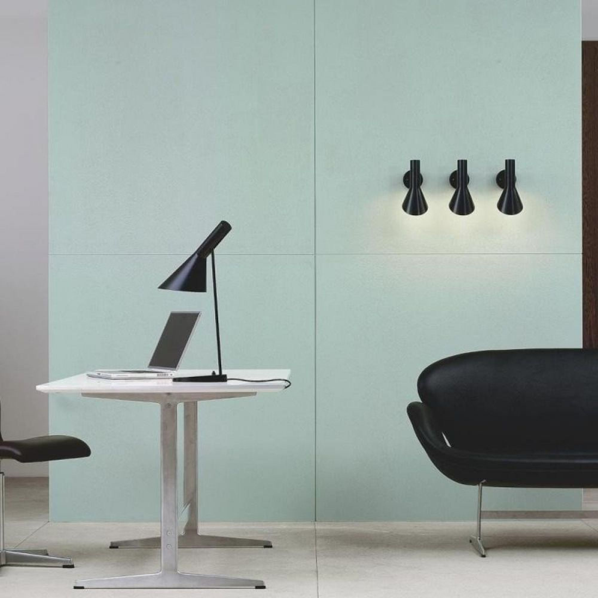 Schreibtisch mit Laptop, schwarzer Lampe, Stuhl und Sofa vor hellgrüner Wand.