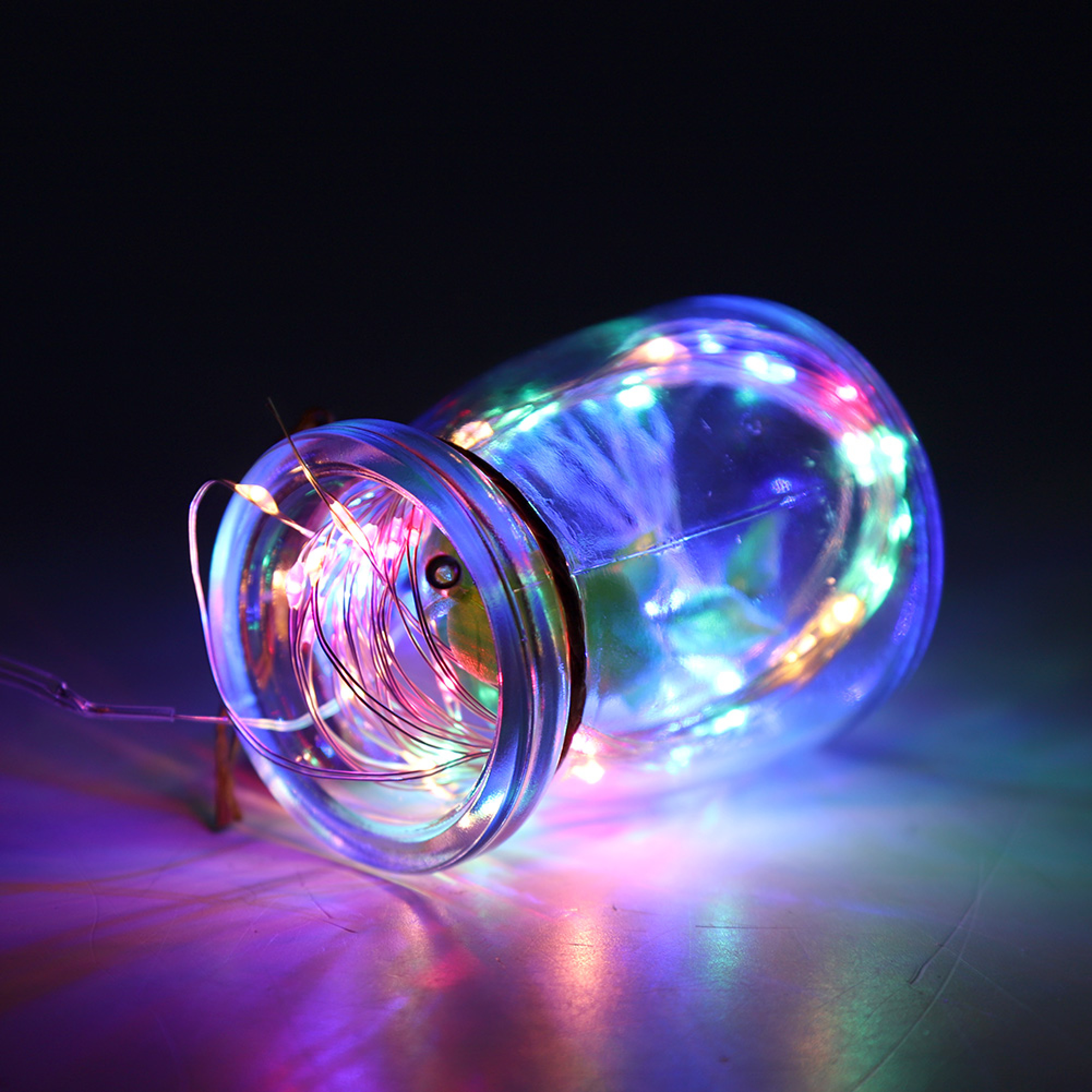 Ein Glasgefäß mit bunten Lichterketten, das auf einer dunklen Fläche liegt.