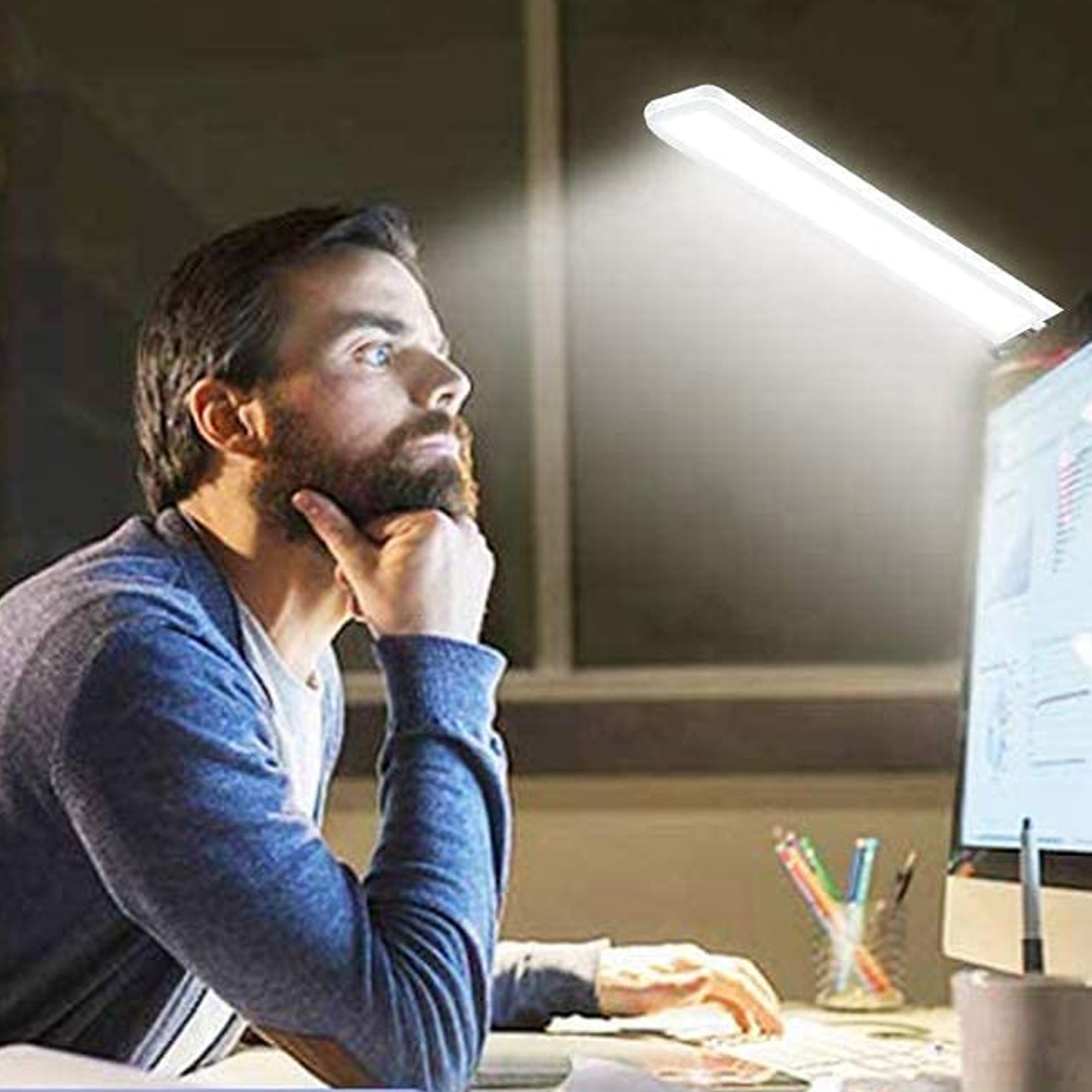 Mann mit Bart blickt auf Computermonitor, beleuchtet von einer hellen LED-Schreibtischlampe.