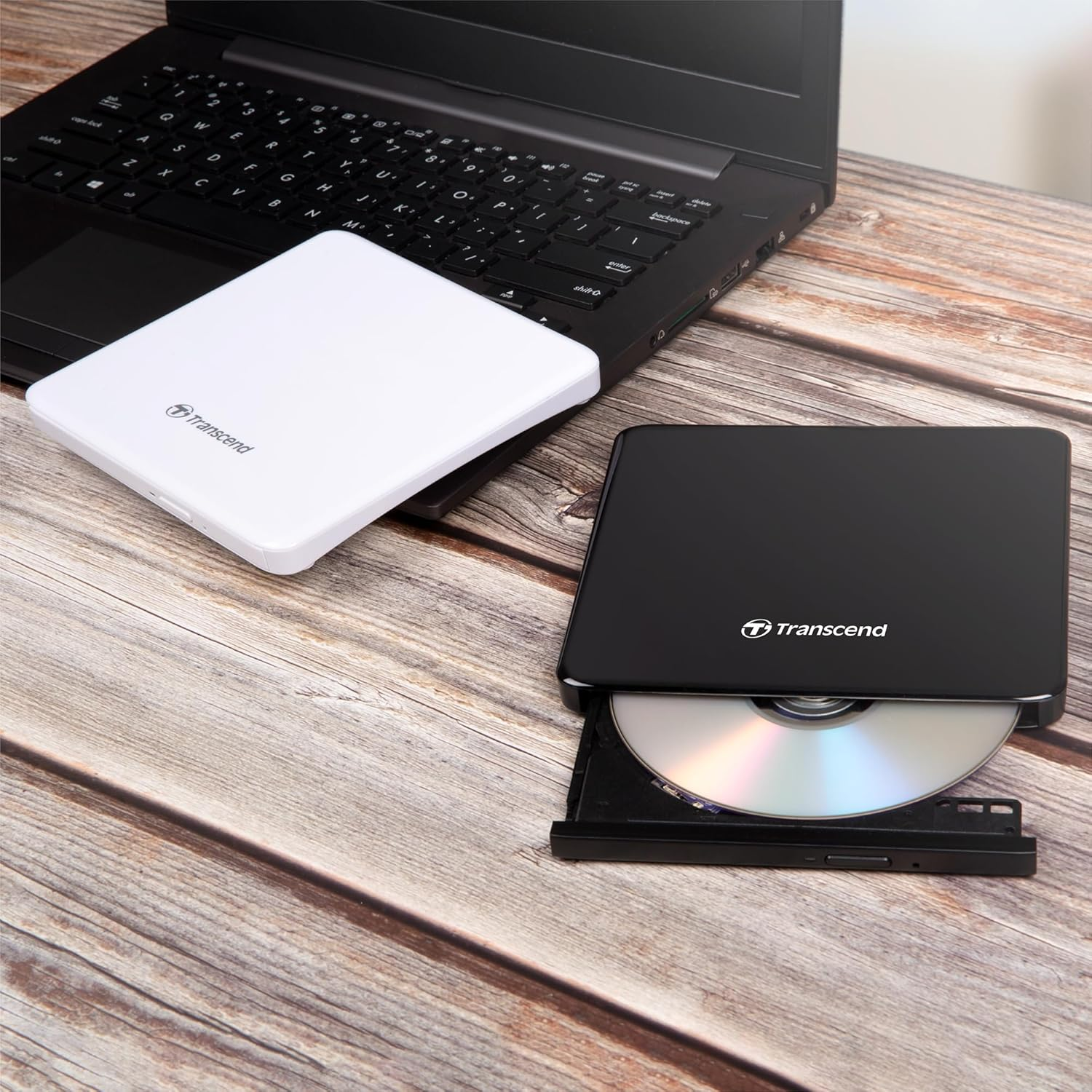 Ein schwarzer Laptop mit einer weißen und einer schwarzen externen CD/DVD-Laufwerk auf Holztisch.