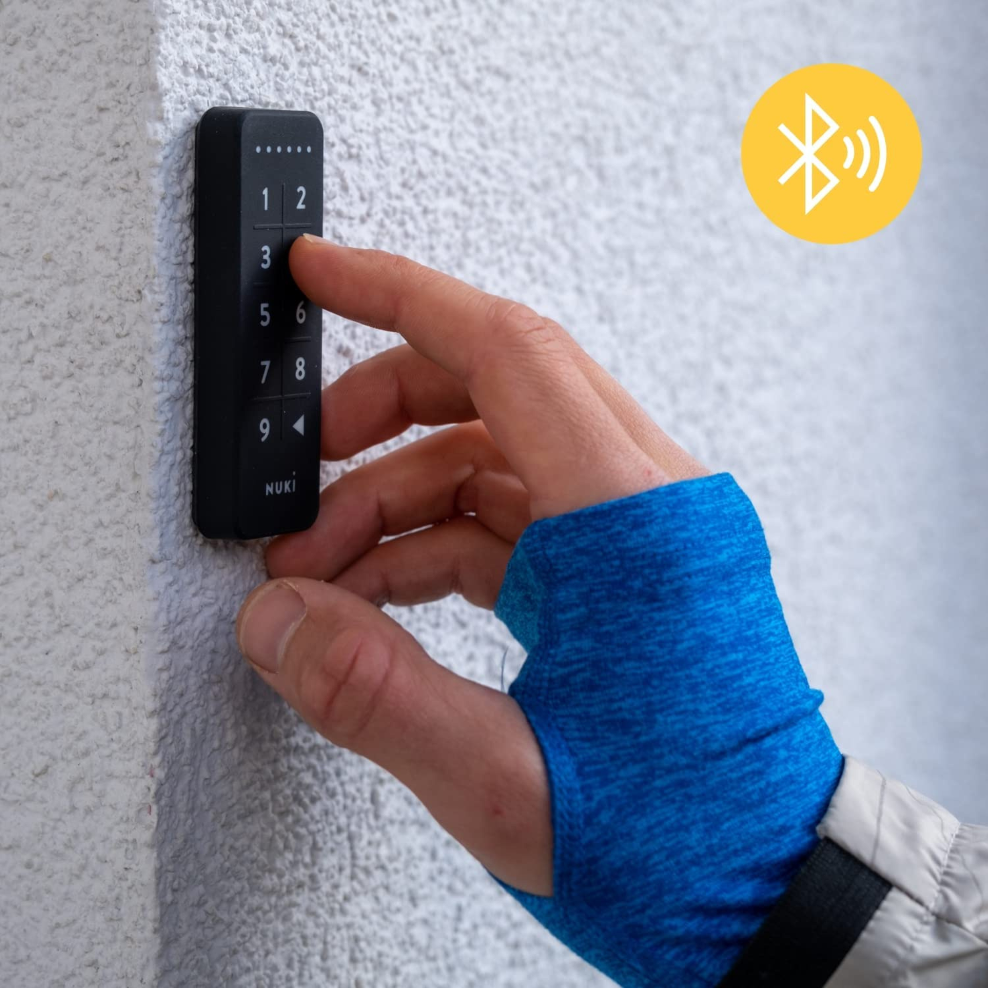 A hand presses a Nuki number keypad mounted on a white wall. A Bluetooth symbol is visible.