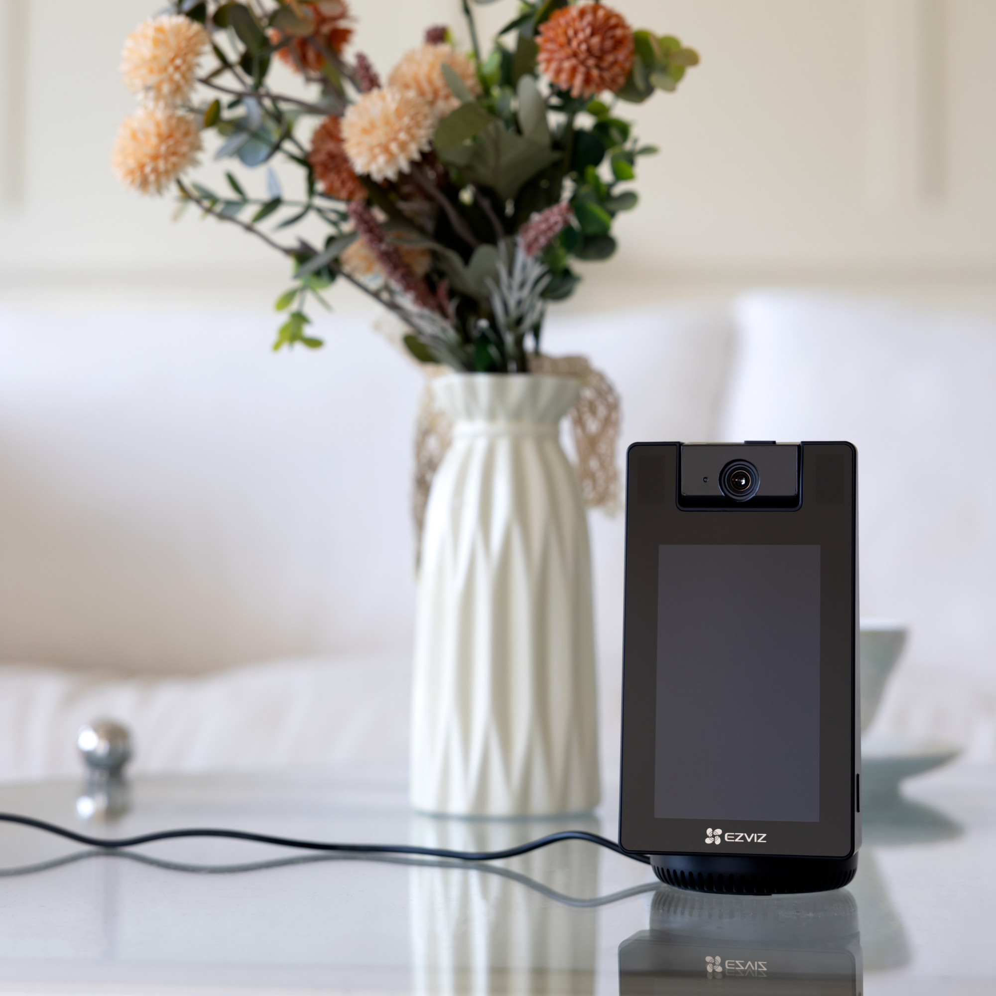 A black smart display sits beside a vase of flowers on a glass table.