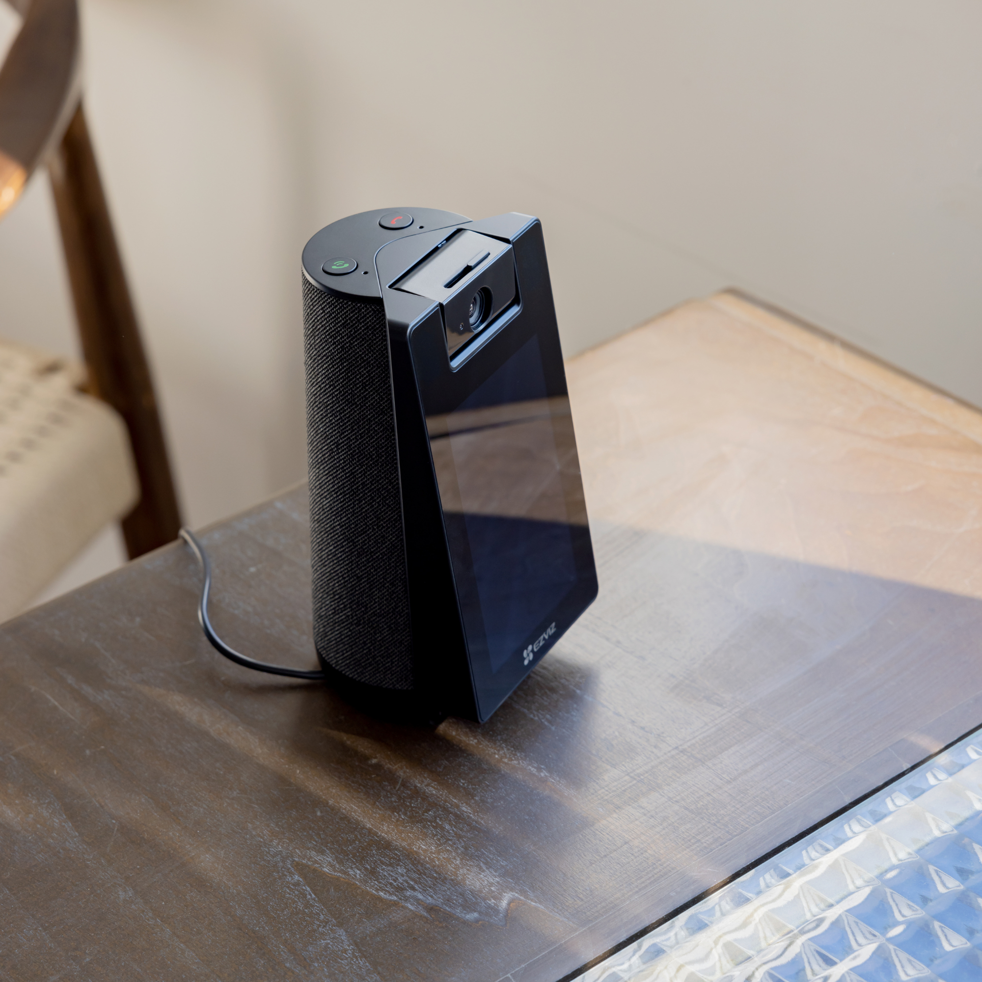 A black smart speaker with a screen is on a wooden table. A chair is visible in the background.