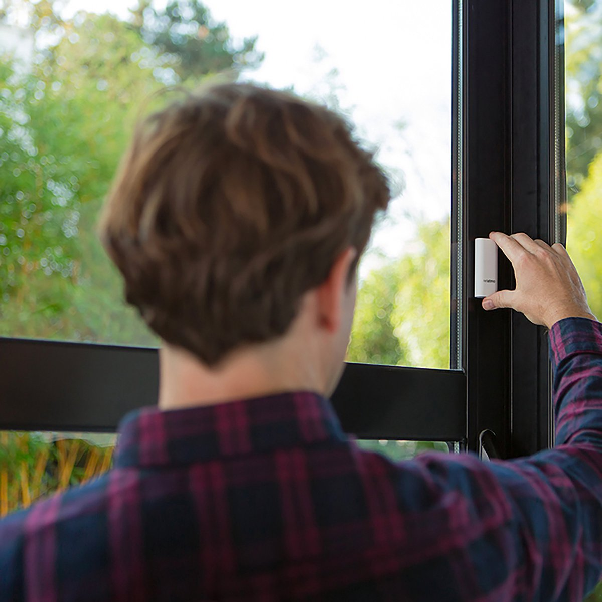 Mężczyzna przyczepia biały czujnik do czarnej ramy okna, widać zielone drzewa.