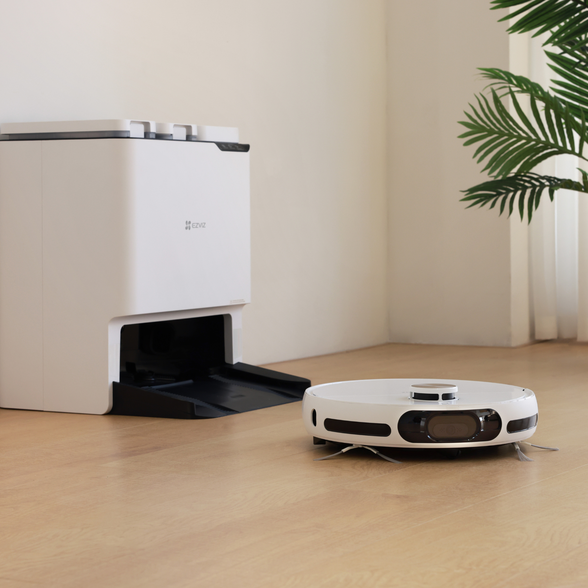 Biały robot sprzątający na drewnianej podłodze obok stacji ładującej z rośliną w tle.