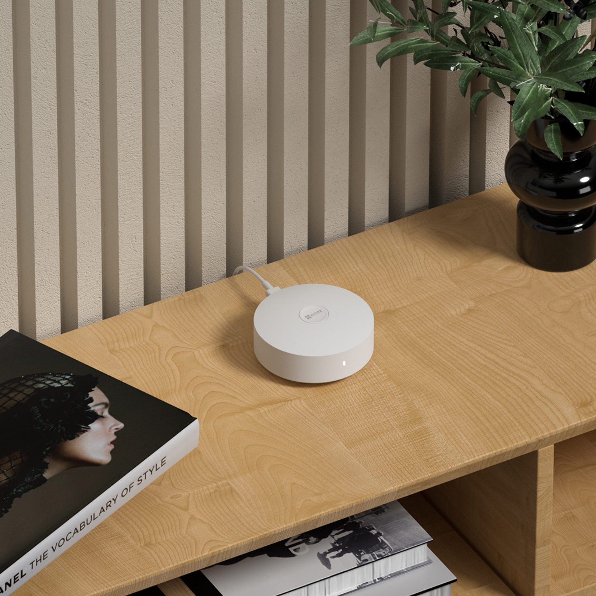 Biały router stoi na drewnianej półce obok książki i czarnego wazonu z zielonymi liśćmi.