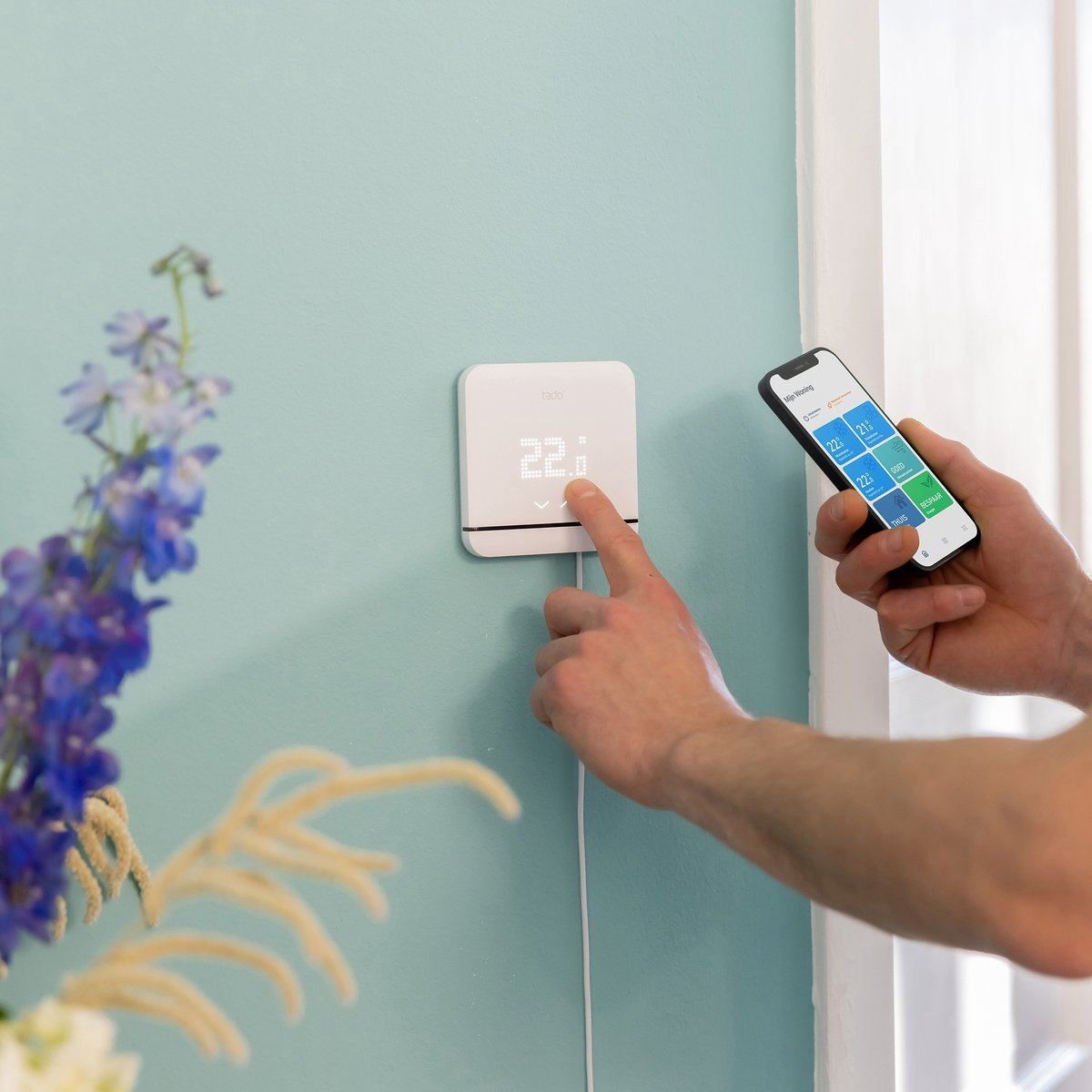 A man is operating a thermostat and holding a smartphone with an app.