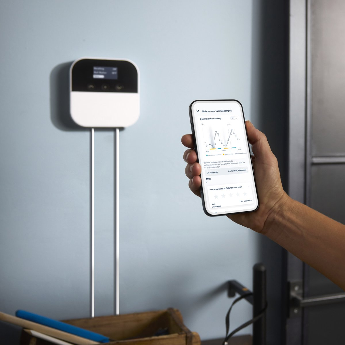 A smartphone shows an app with a graph next to a white heating controller on the wall.