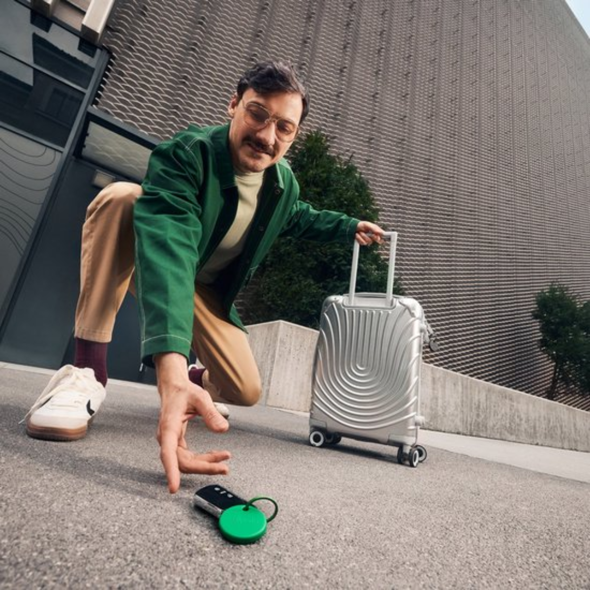 A man in a green jacket and tan pants kneels, reaching for keys with a green tracker on the ground next to a silver suitcase.