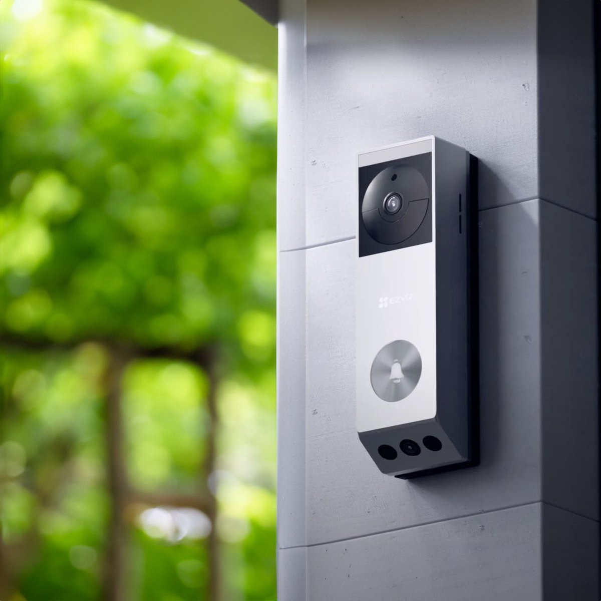 A silver video doorbell with a camera, bell icon, and three sensors on a wall.