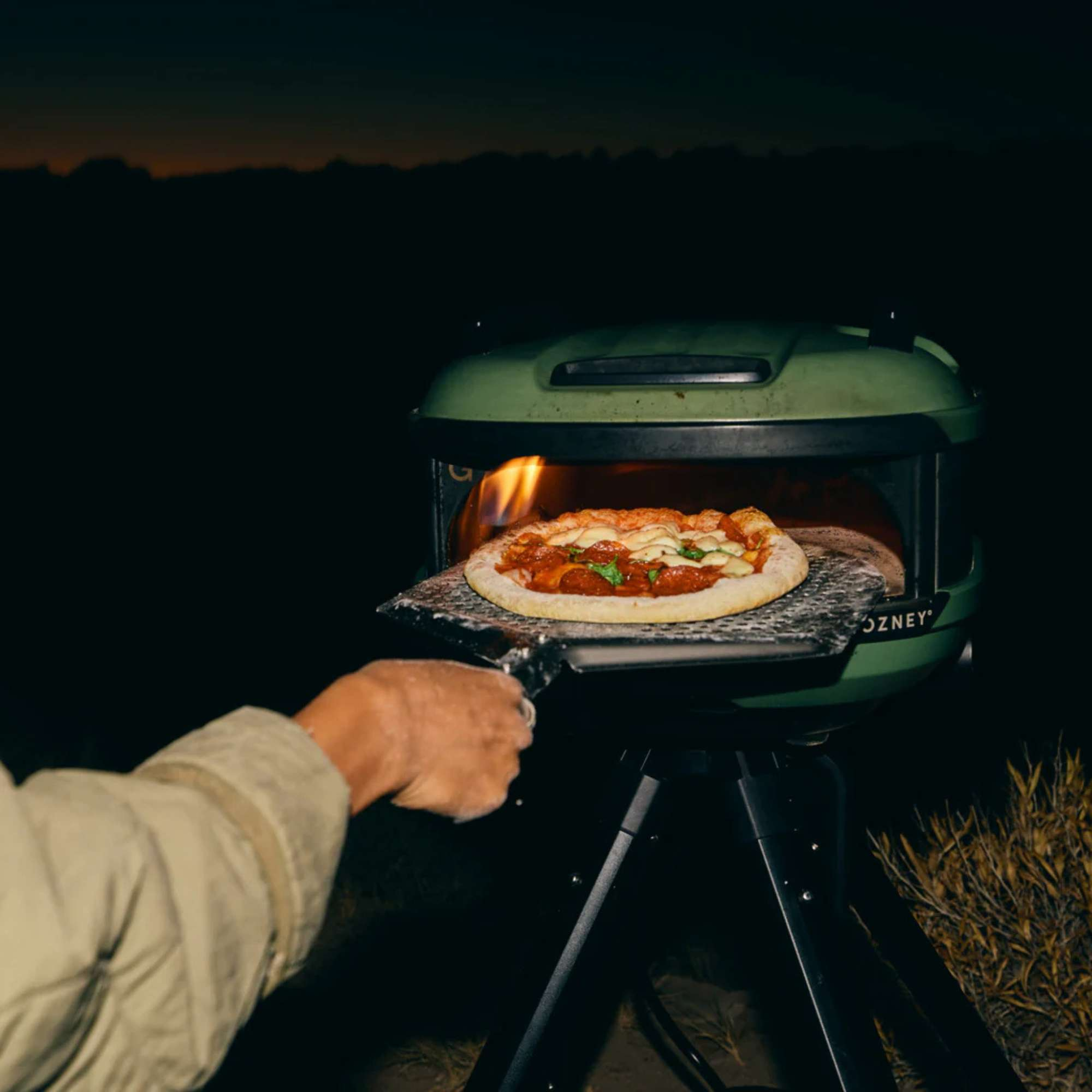 Eine Person kocht eine Peperoni-Pizza in einem grünen Pizzaofen im Freien in der Dämmerung.