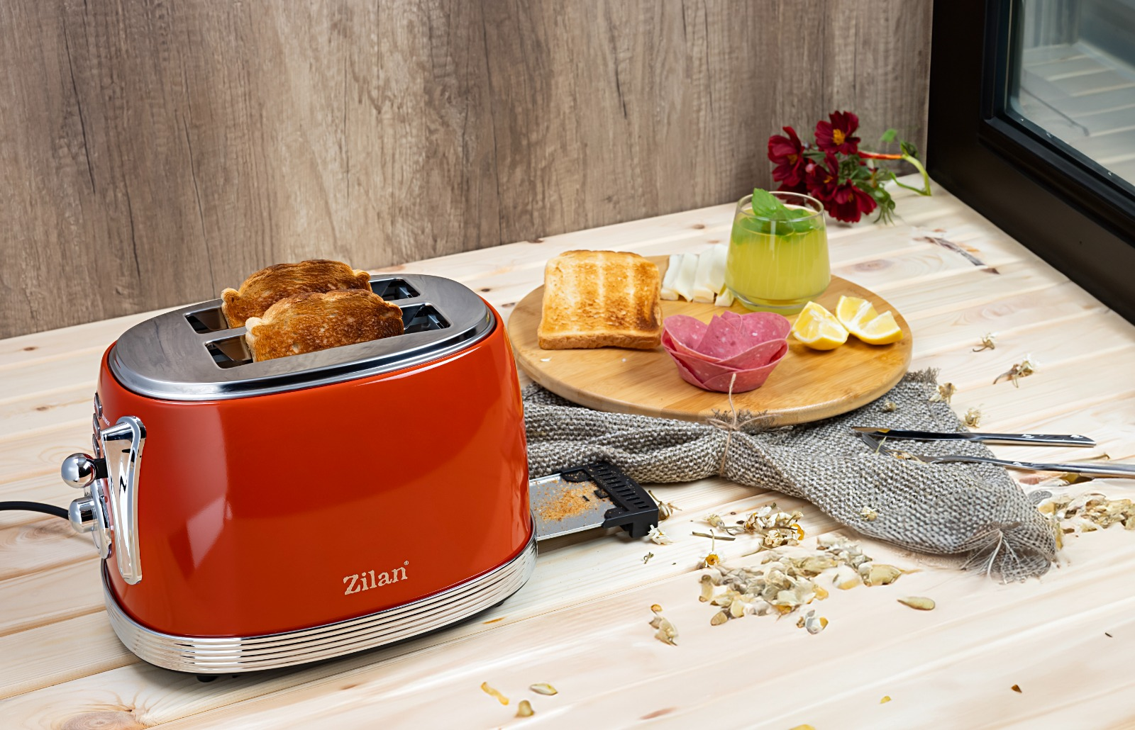 Ahşap bir masada, kızarmış ekmekli kırmızı bir tost makinesi, bir bardak meyve suyu, limon ve sosis.