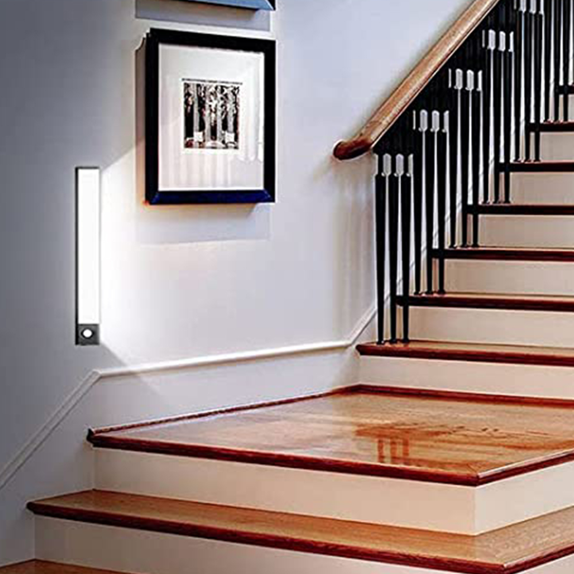 Ein langer, schmaler Wandleuchter beleuchtet eine Wand neben einer Holztreppe mit schwarzen Geländern.
