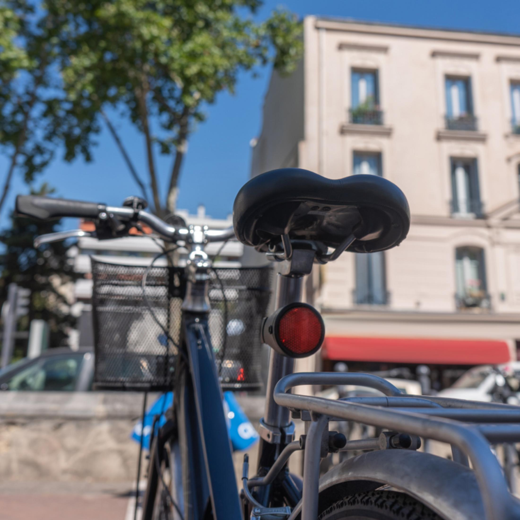 Fahrradsattel mit rotem Reflektor, Korb und Gepäckträger, Gebäude im Hintergrund.
