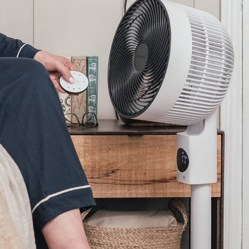 Eine Person in dunkelblauer Kleidung bedient einen weißen Ventilator mit Fernbedienung.