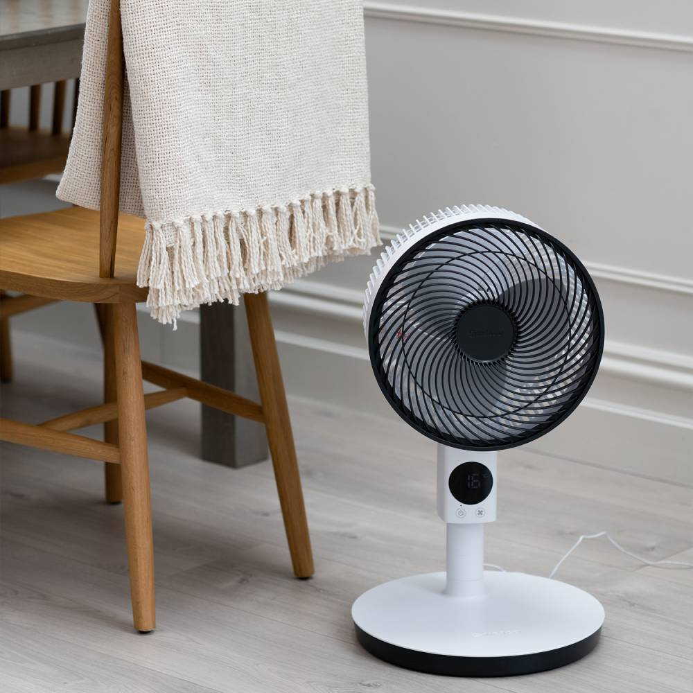 Weißer Ventilator mit schwarzem Gitter neben einem Holzstuhl mit cremefarbener Decke.