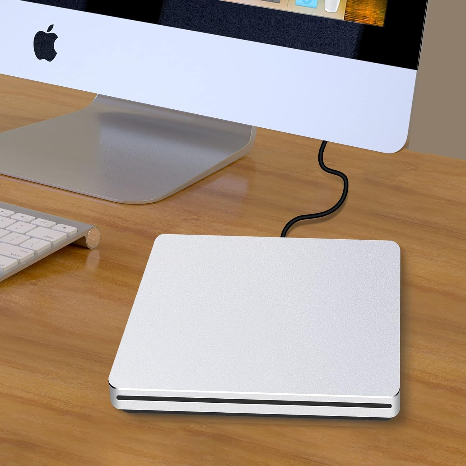 Ein silbernes externes CD/DVD-Laufwerk liegt auf einem Holztisch neben einer Tastatur und einem iMac.