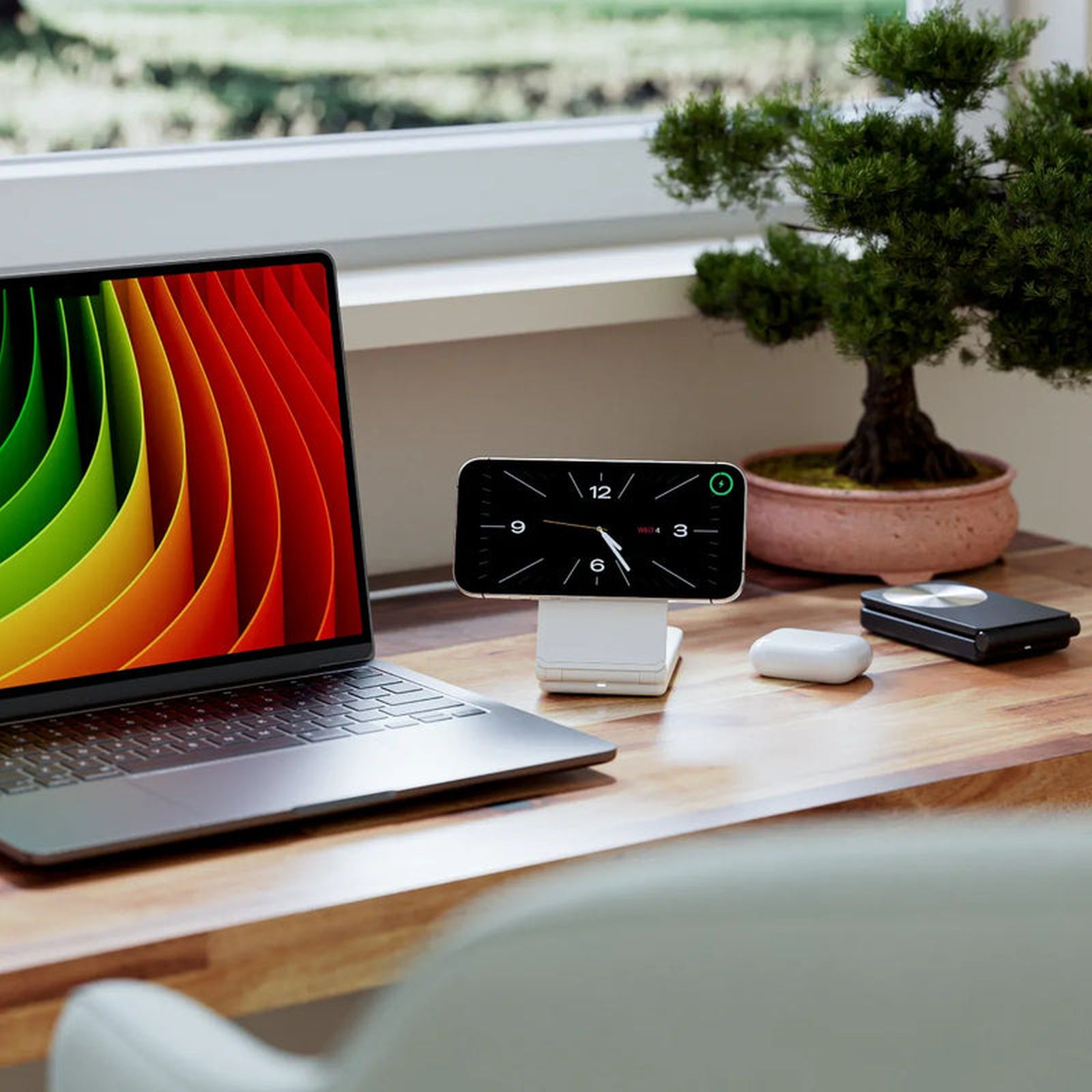 Ein Laptop mit buntem Hintergrund, ein Handy auf Ladestation, Airpods und ein Bonsai.