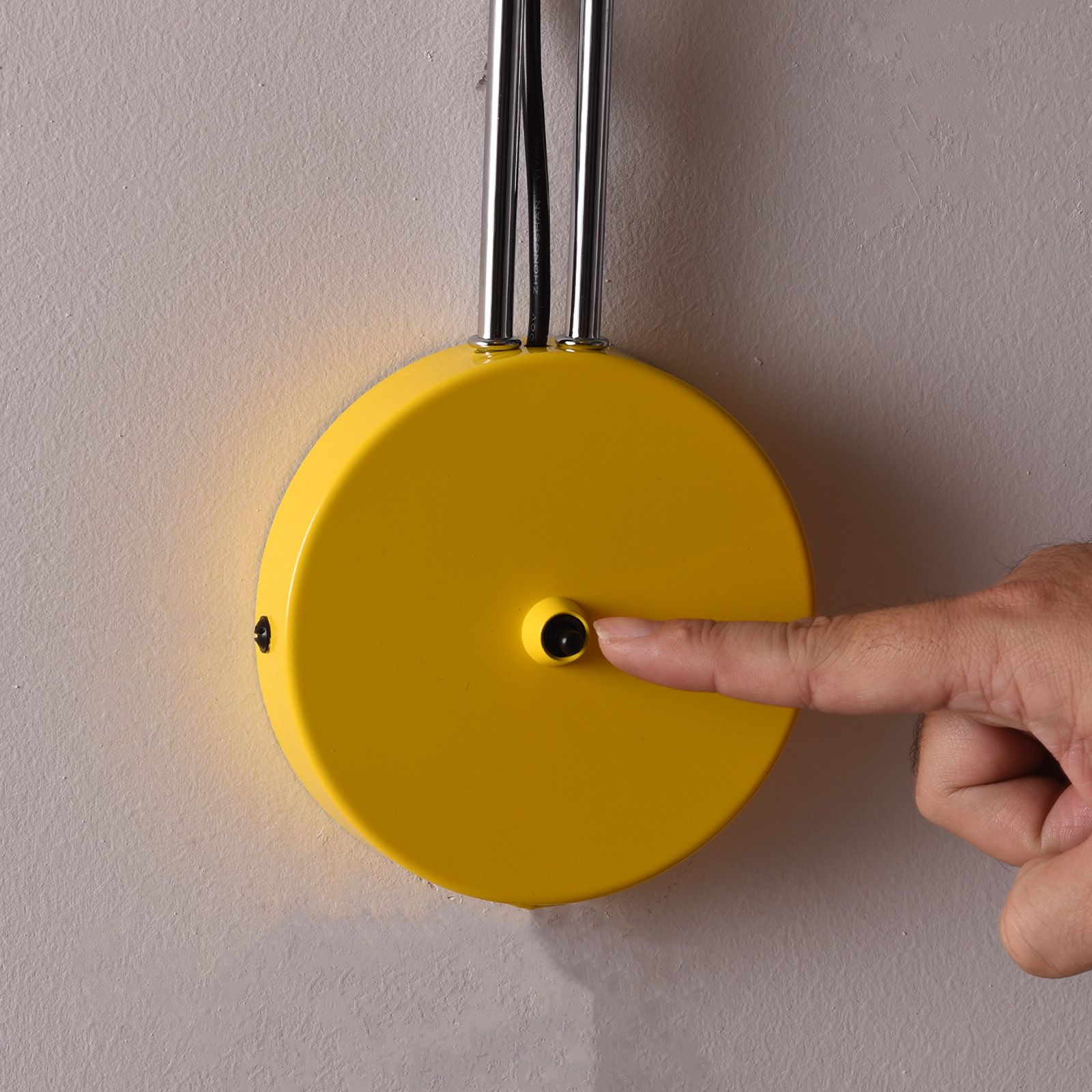 Un dedo presiona un interruptor de luz circular amarillo en una pared gris.