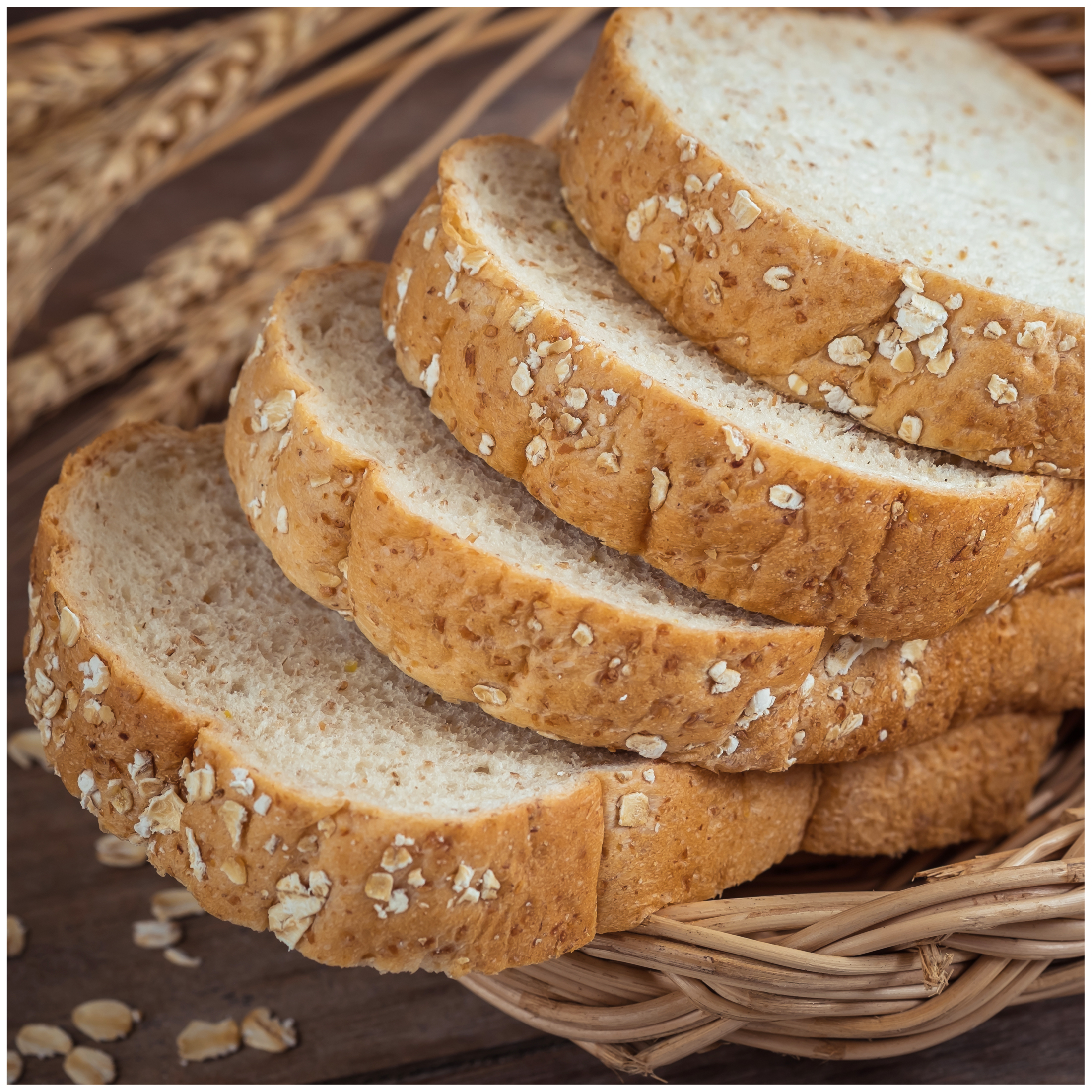 Fette di pane all'avena impilate in un cesto con spighe di grano sullo sfondo.