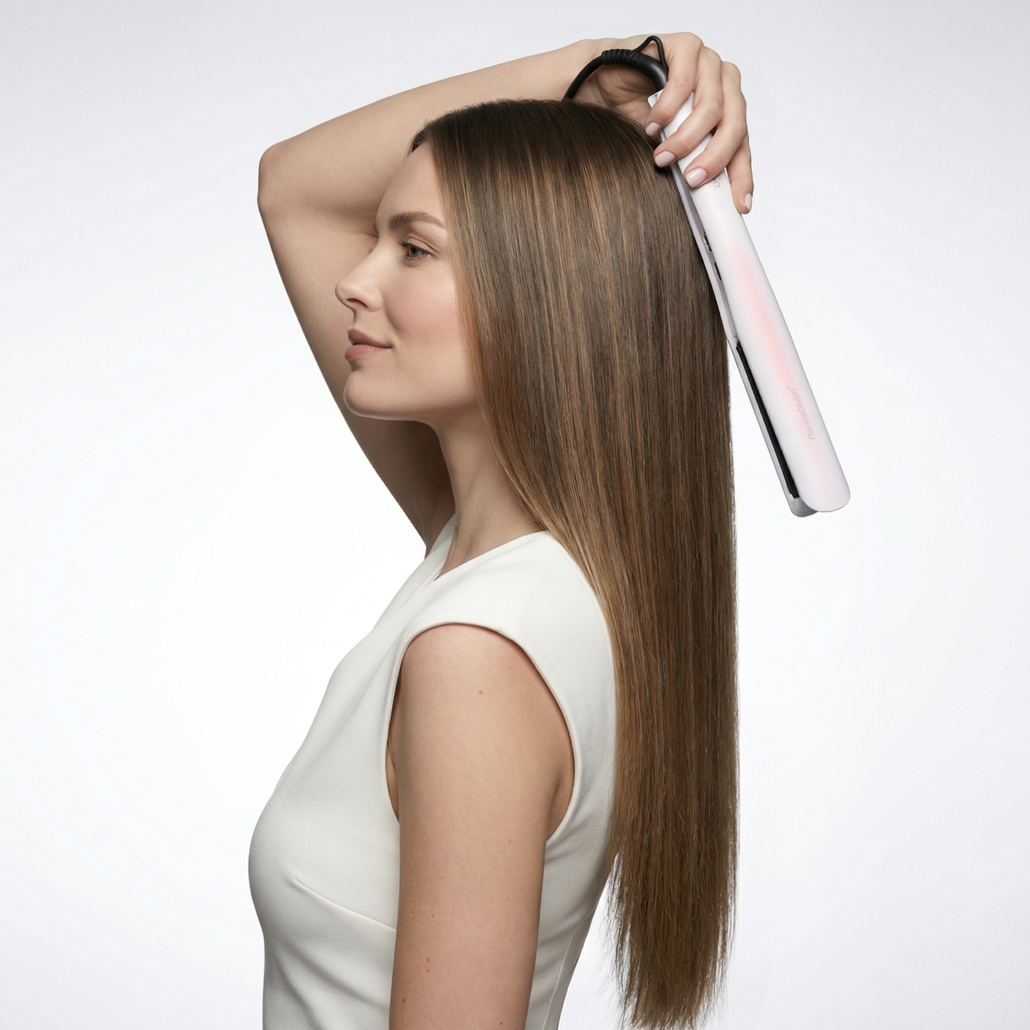 Woman in white dress uses a white hair straightener on her long brown hair.