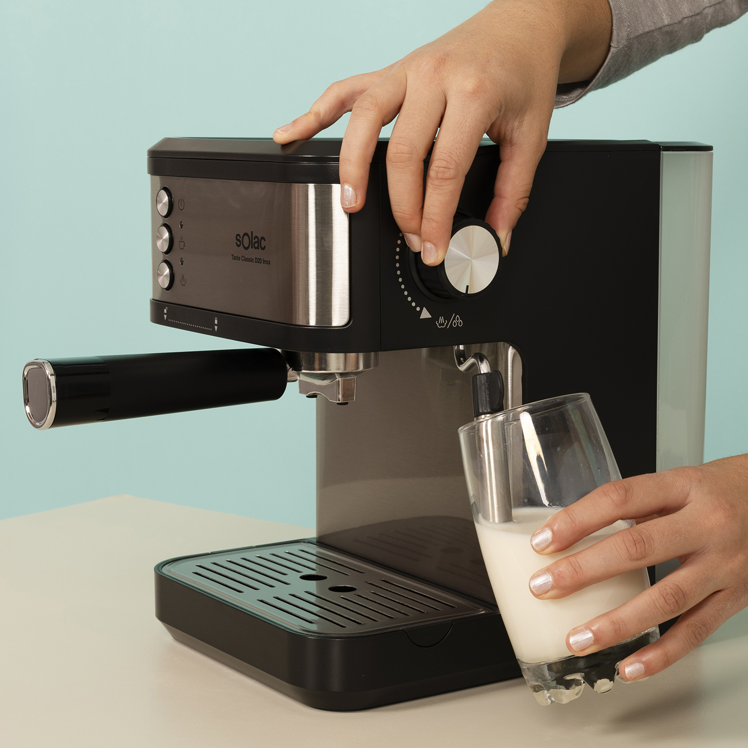 Manos operan una cafetera negra, una ajusta un pomo, la otra sostiene un vaso de leche.