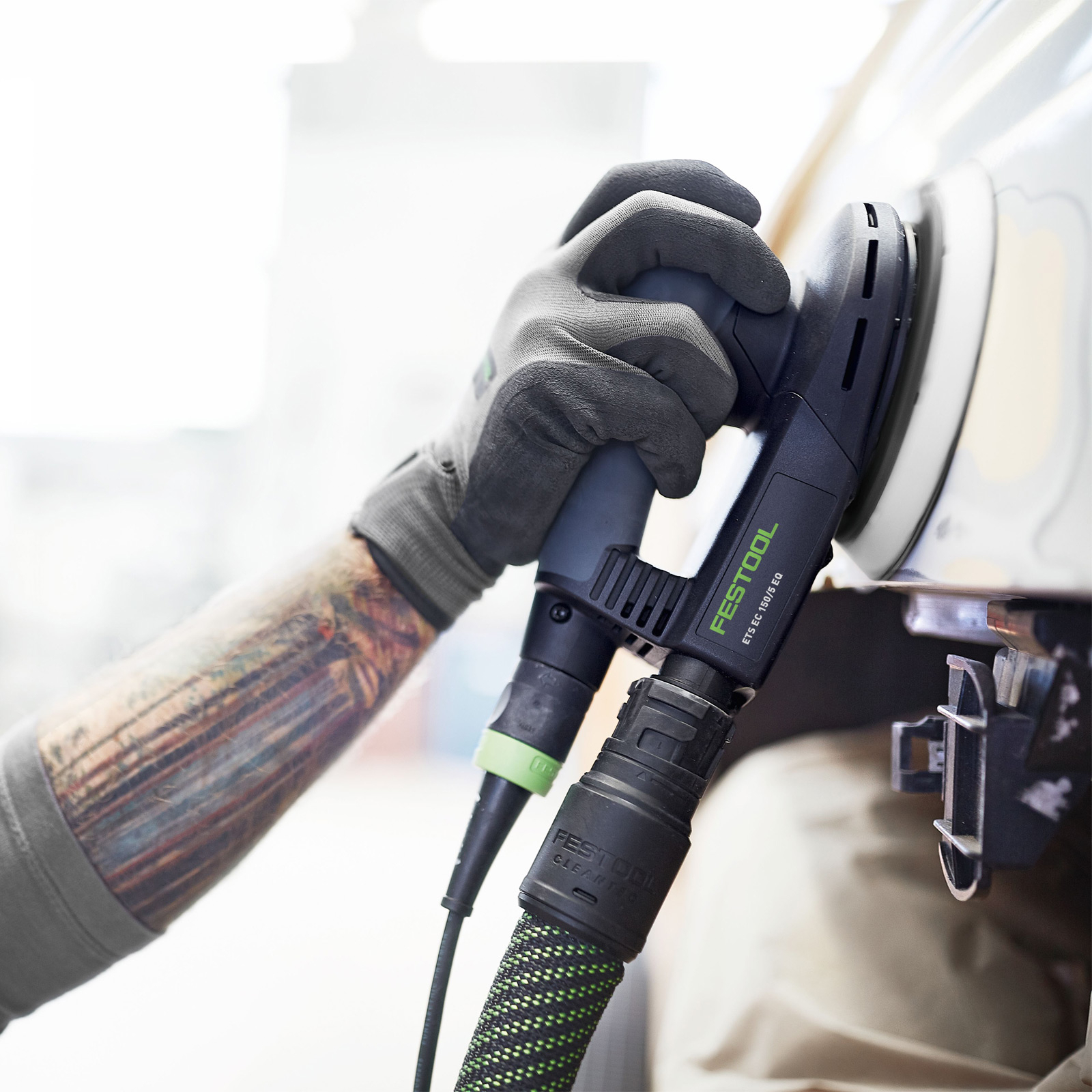 Hand mit Handschuh hält Schleifmaschine an Auto. Tattoos am Arm sichtbar.