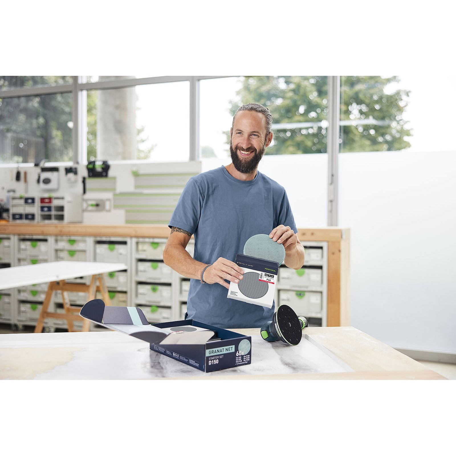 Ein Mann mit Bart in einem blauen T-Shirt hält Schleifpapier in einer Werkstatt.