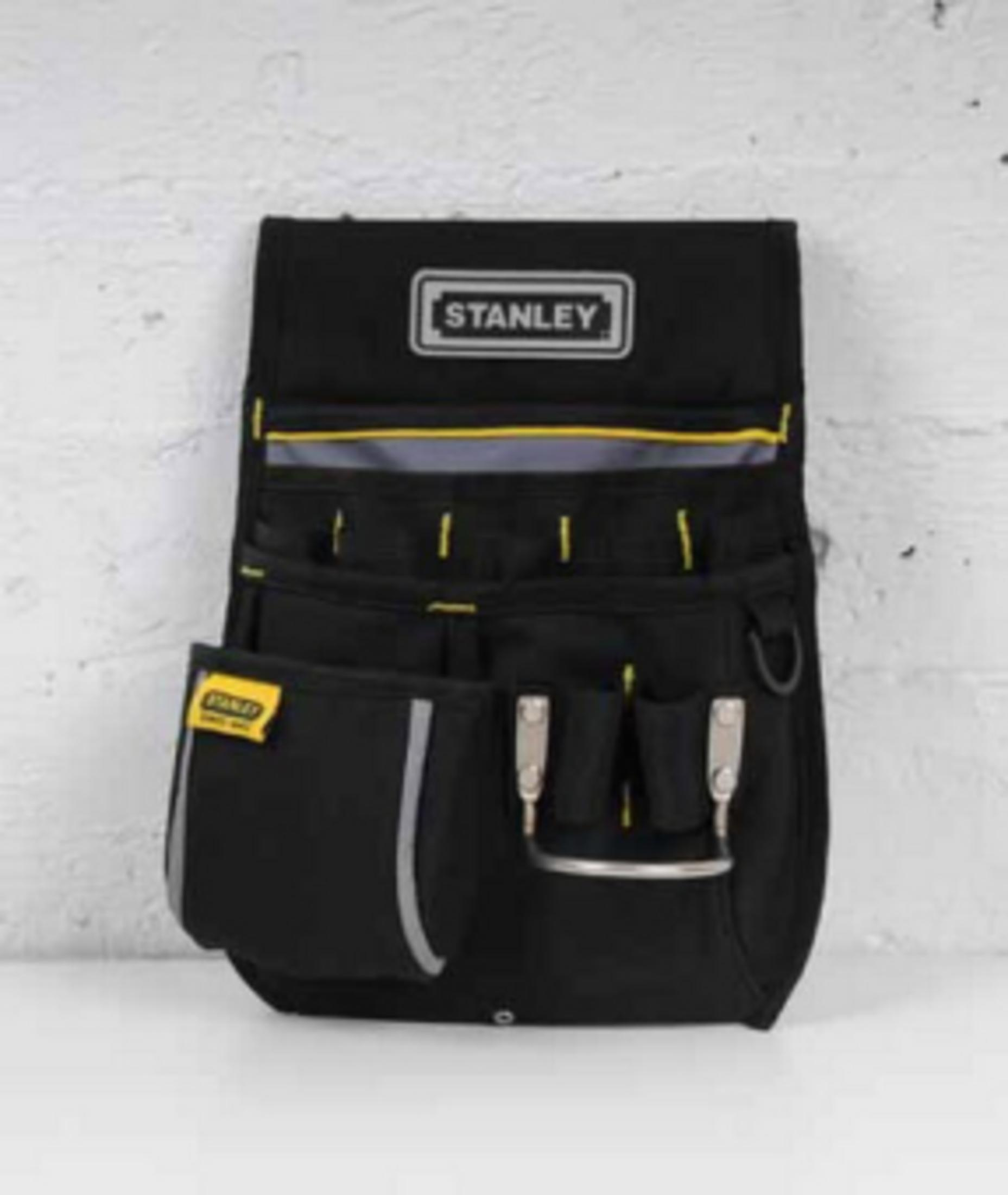 Marsupio porta attrezzi nero con logo Stanley e dettagli gialli, su sfondo di mattoni.