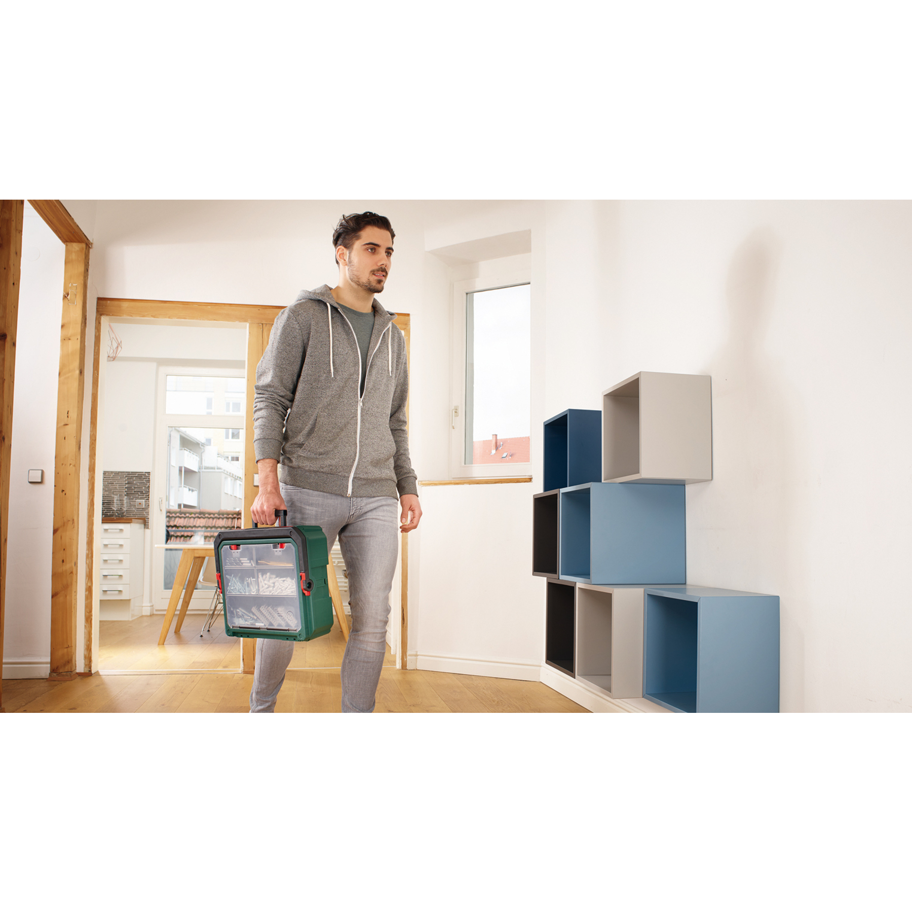 Mann in grauem Hoodie trägt Werkzeugkasten durch einen Raum mit Holzdetails.