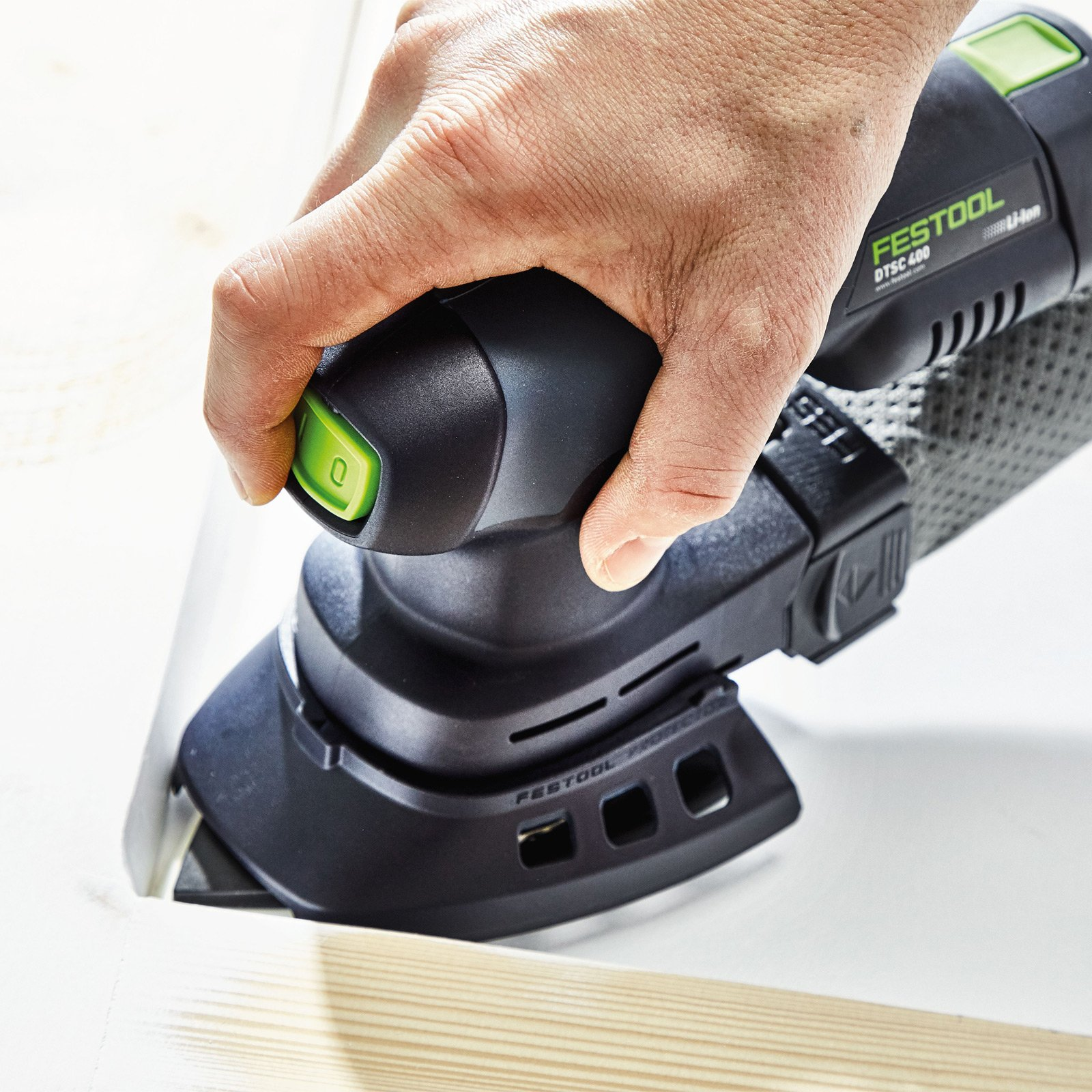 Eine Hand drückt den grünen Knopf an einem schwarzen Festool-Schleifer, der an Holz arbeitet.