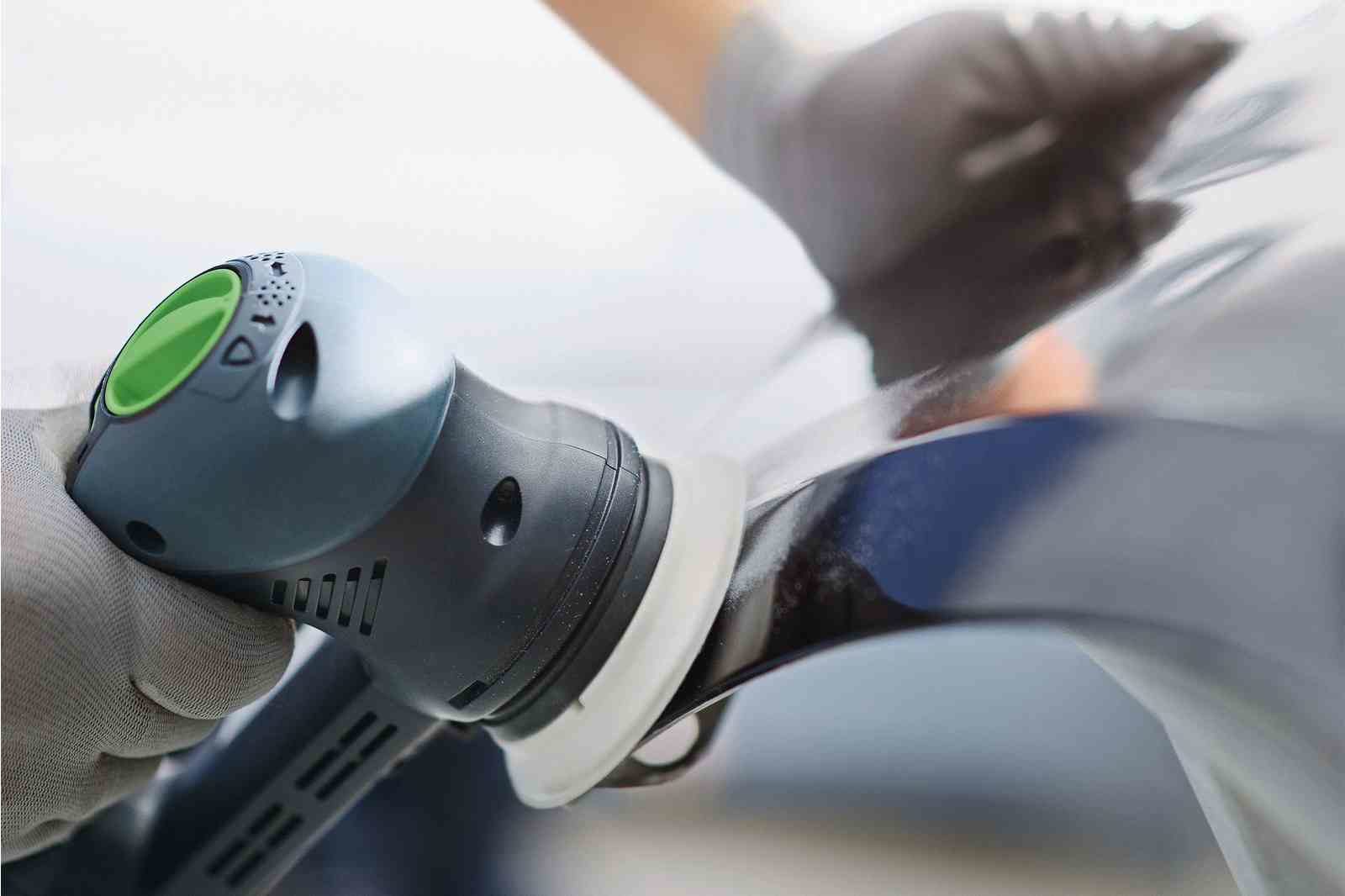 Eine behandschuhte Hand poliert eine glänzende Oberfläche mit einem grauen und grünen Exzenterschleifer.