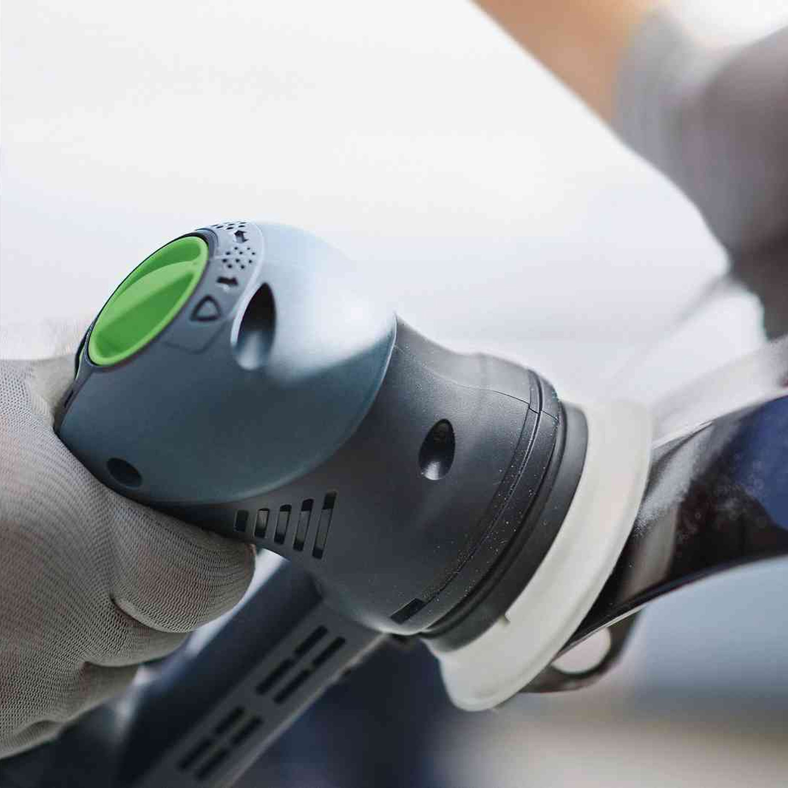 Grauer Exzenterschleifer wird von einer Hand mit Handschuh auf einer glatten Oberfläche gehalten.