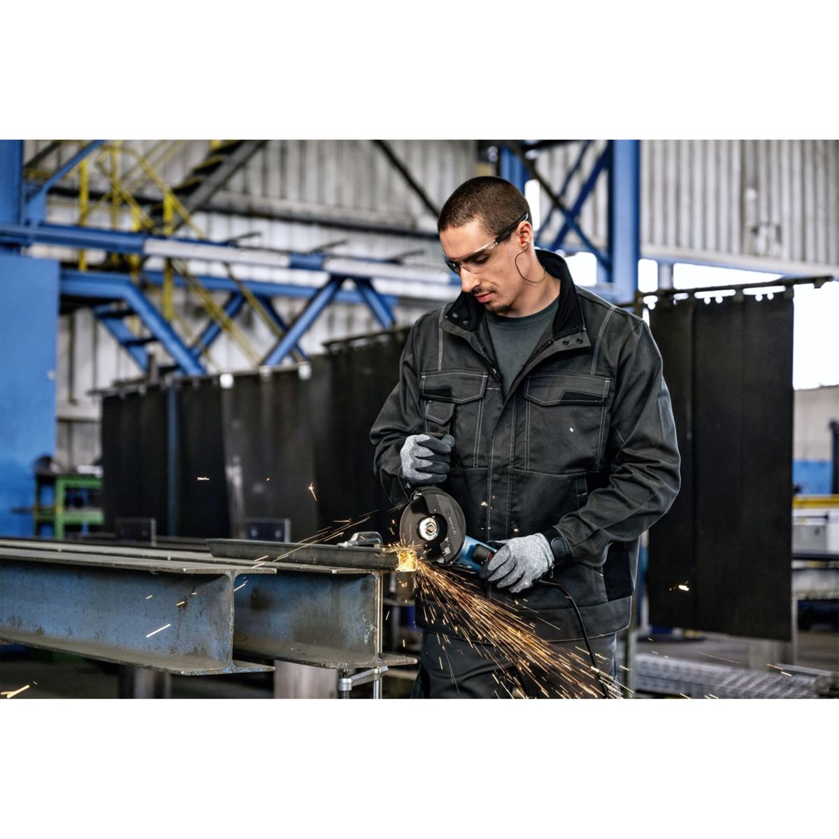 Ein Mann in Schutzkleidung benutzt einen Winkelschleifer an Metallträgern.