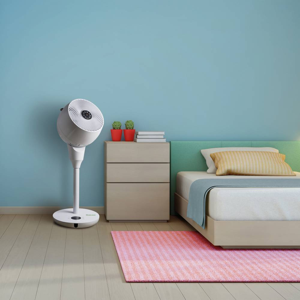Un ventilador blanco de pie está junto a una mesita de noche con dos plantas pequeñas y libros.