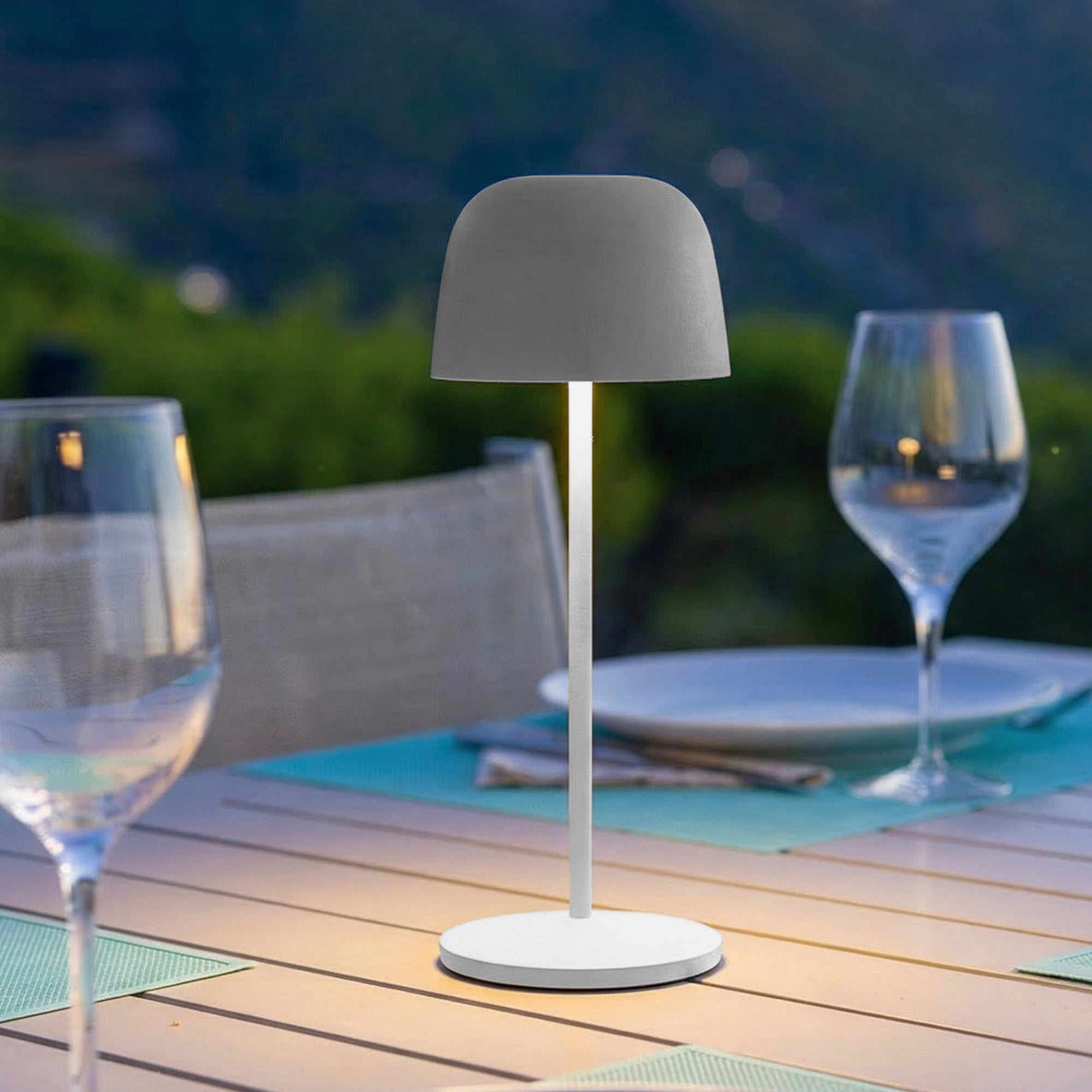 Graue Tischlampe auf einem Tisch mit Gläsern und Tellern bei Dämmerung.