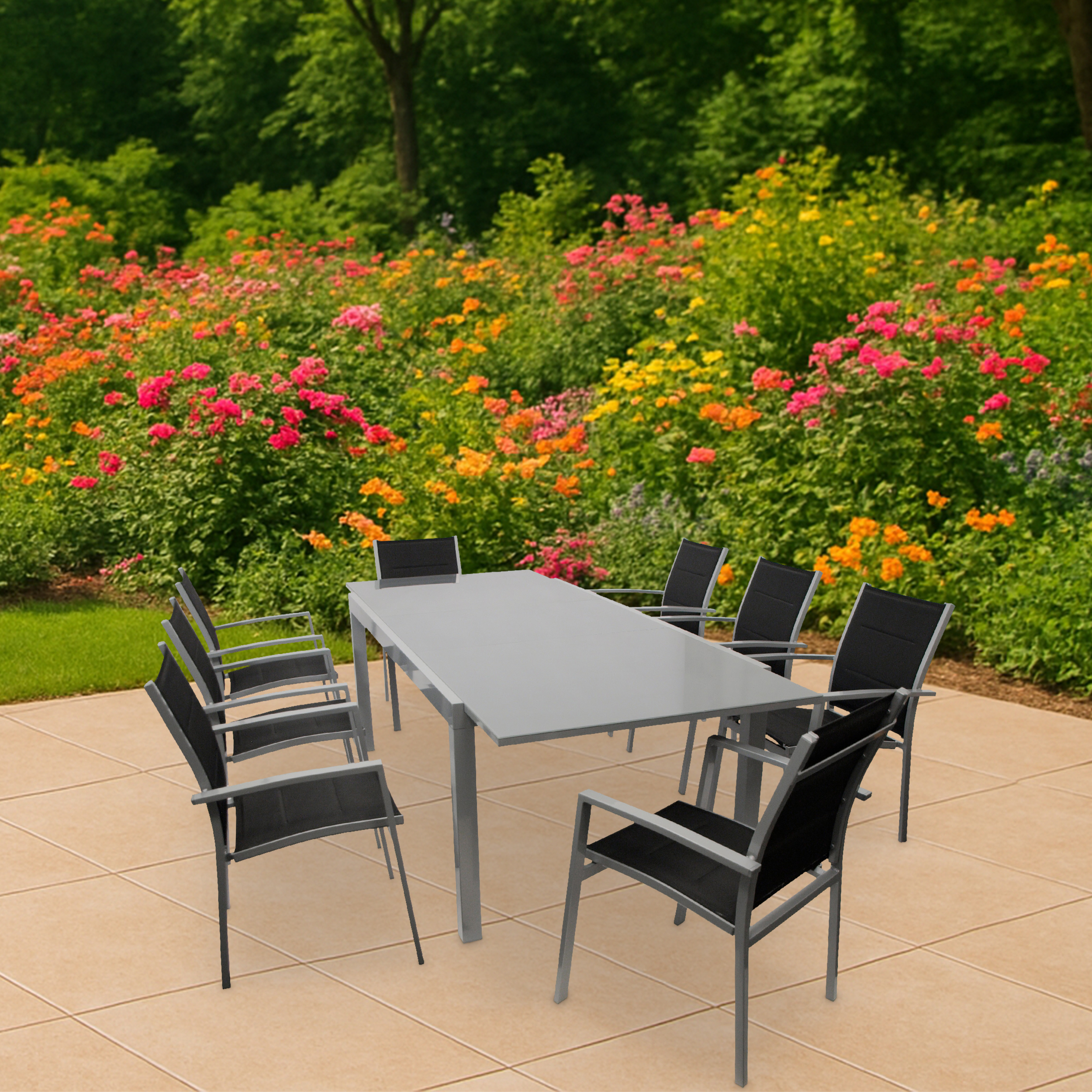 Ein grauer Tisch mit schwarzen Stühlen steht auf einer hellbraunen Terrasse vor blühenden Blumen.
