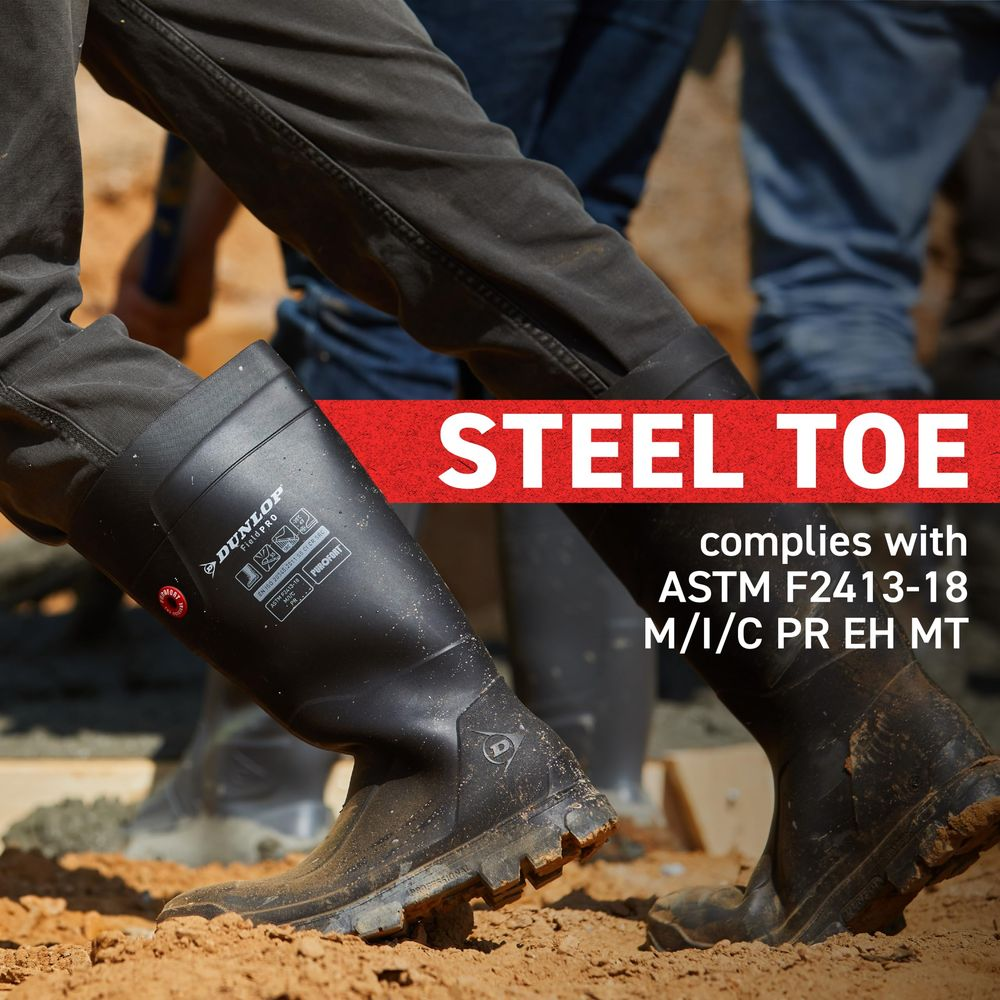 Schwarze Gummistiefel mit Stahlkappe, die auf sandigem Untergrund stehen.