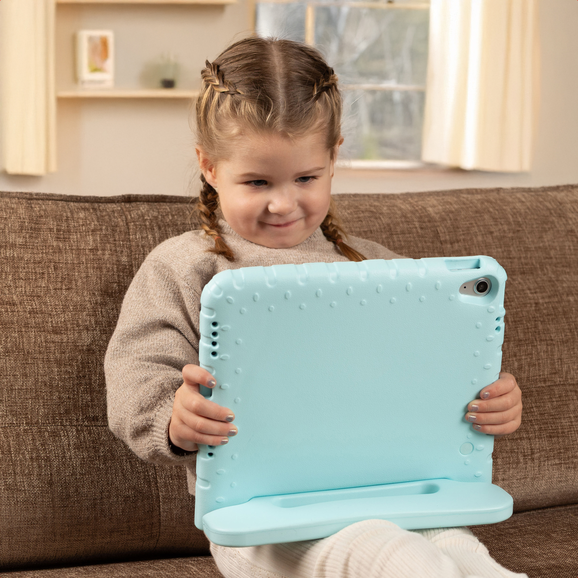 Une jeune fille aux cheveux tressés est assise sur un canapé marron tenant une tablette bleu clair.