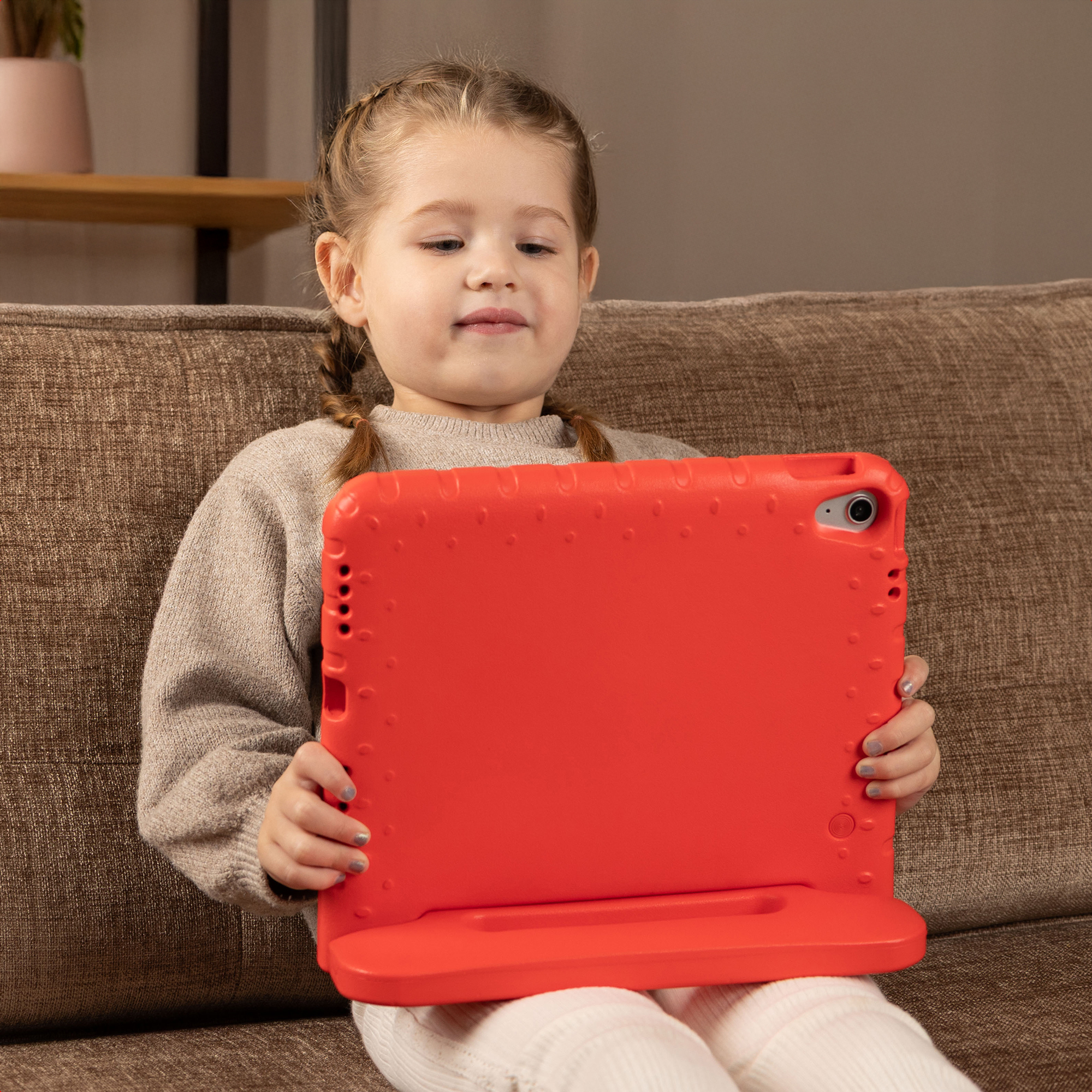 Une petite fille avec des tresses est assise sur un canapé marron, tenant une tablette rouge.